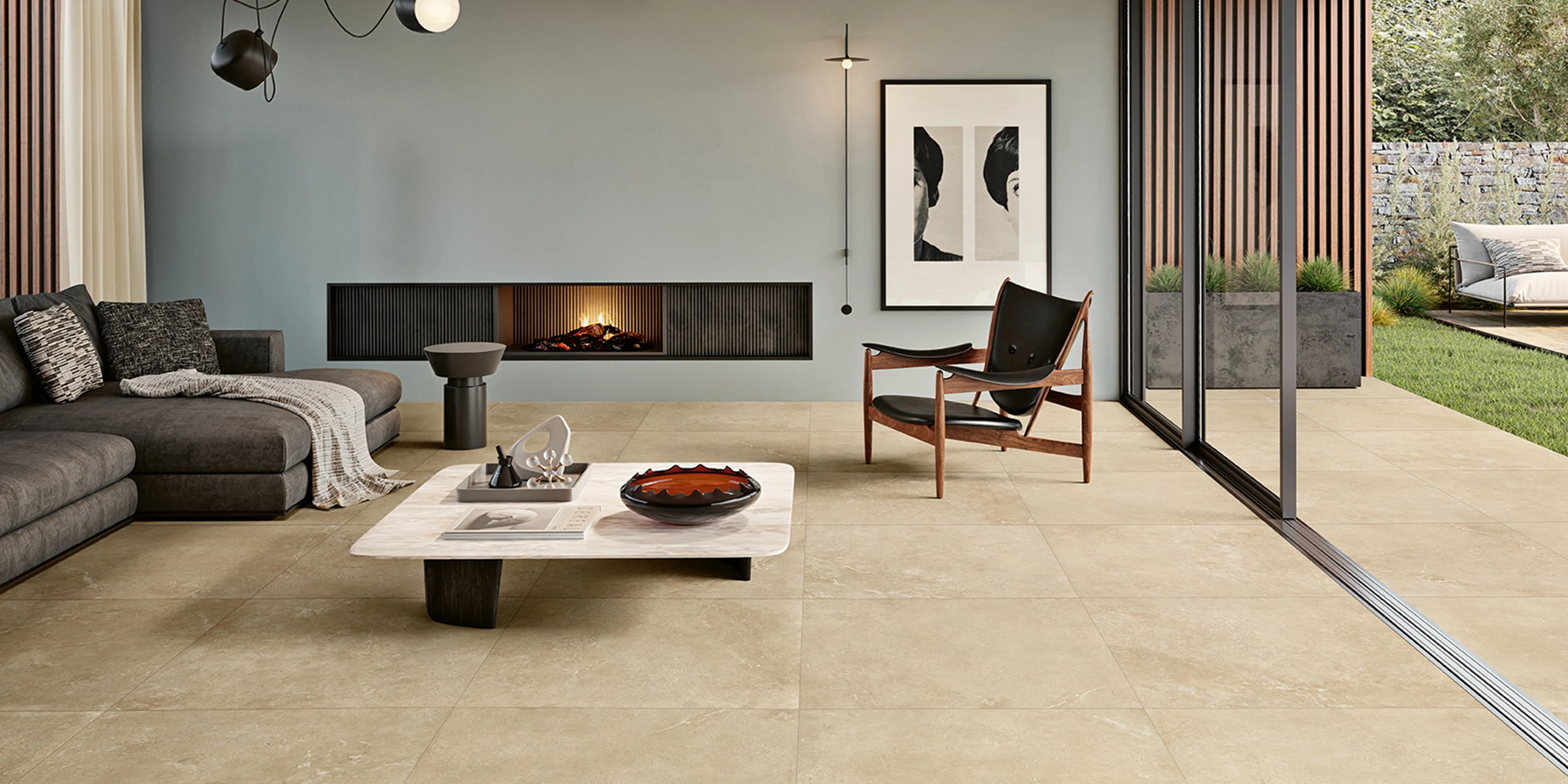 Modern living room with beige tile flooring, gray sectional sofa, fireplace, and sliding glass doors to a garden.