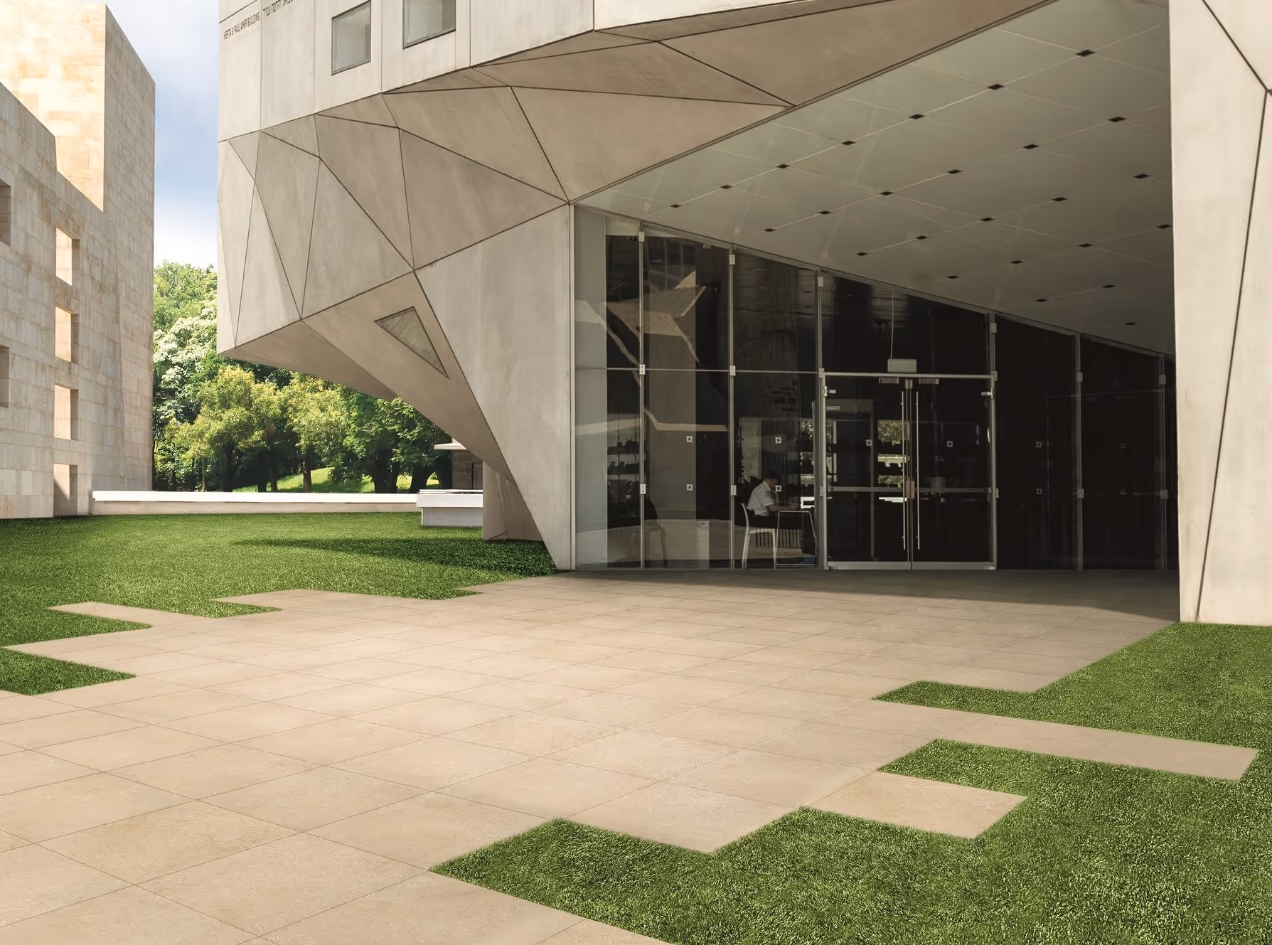 Modern building entrance with geometric concrete facade, glass doors, tiled area, and surrounding green grass.