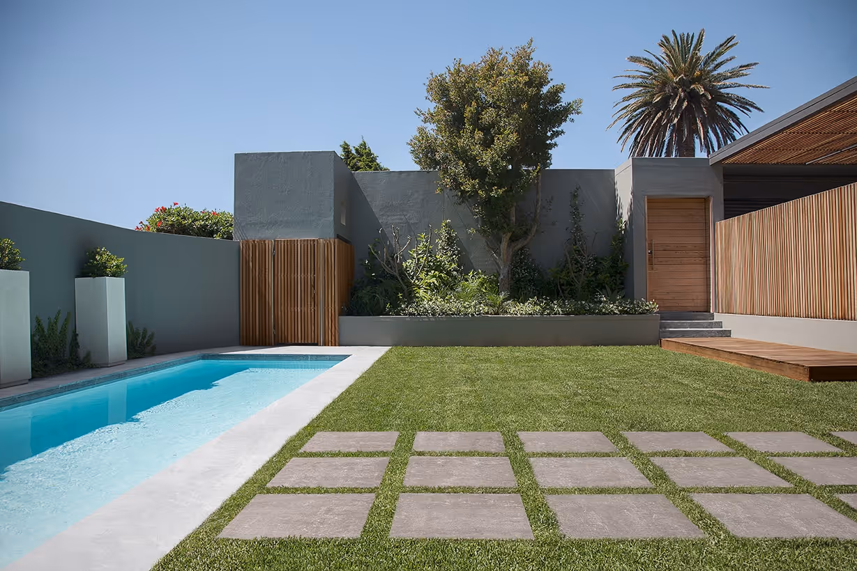 Modern backyard with rectangular pool, stone path on grass, wooden doors, and trees against a clear sky.