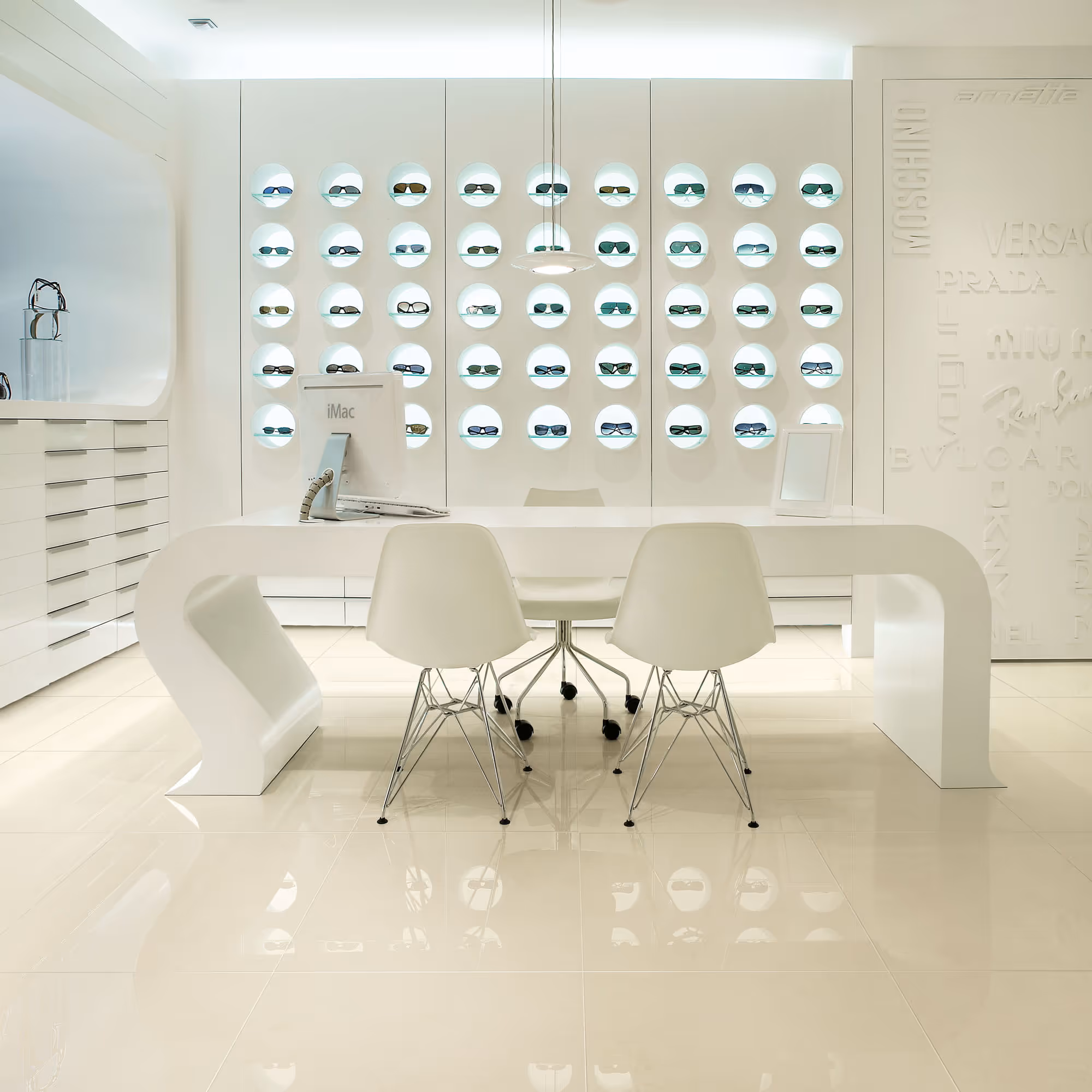 Modern eyewear store interior with white table, chairs, iMac, and sunglasses displayed in lit wall niches.