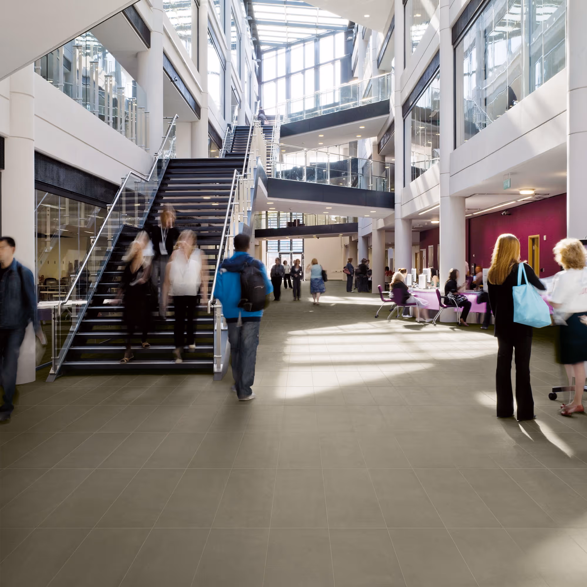 Bright modern lobby with glass staircases and people walking, talking, and sitting at tables.