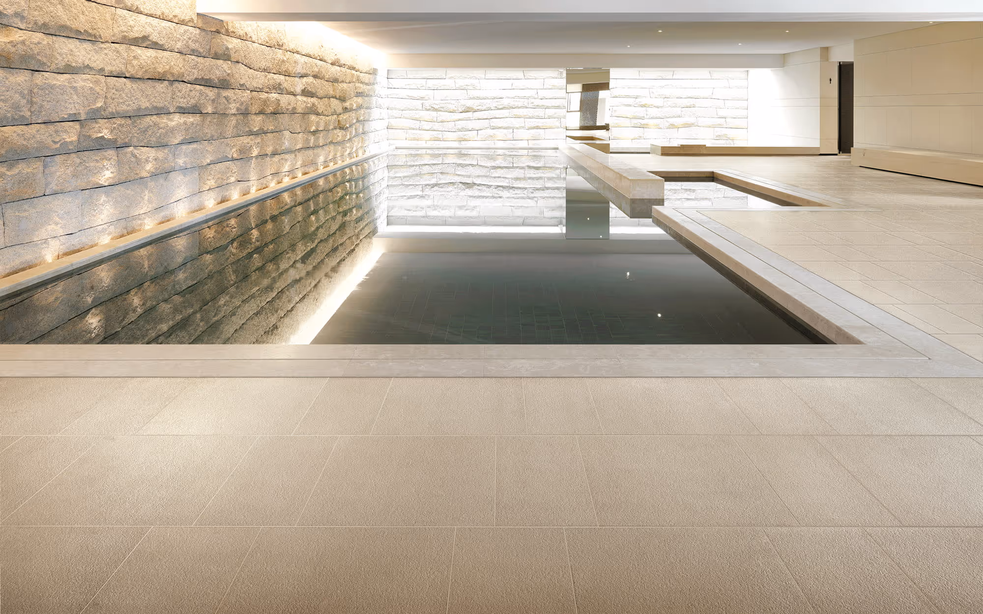 Indoor modern pool with stone walls, reflective water, beige tiled floor, and ceiling lights.