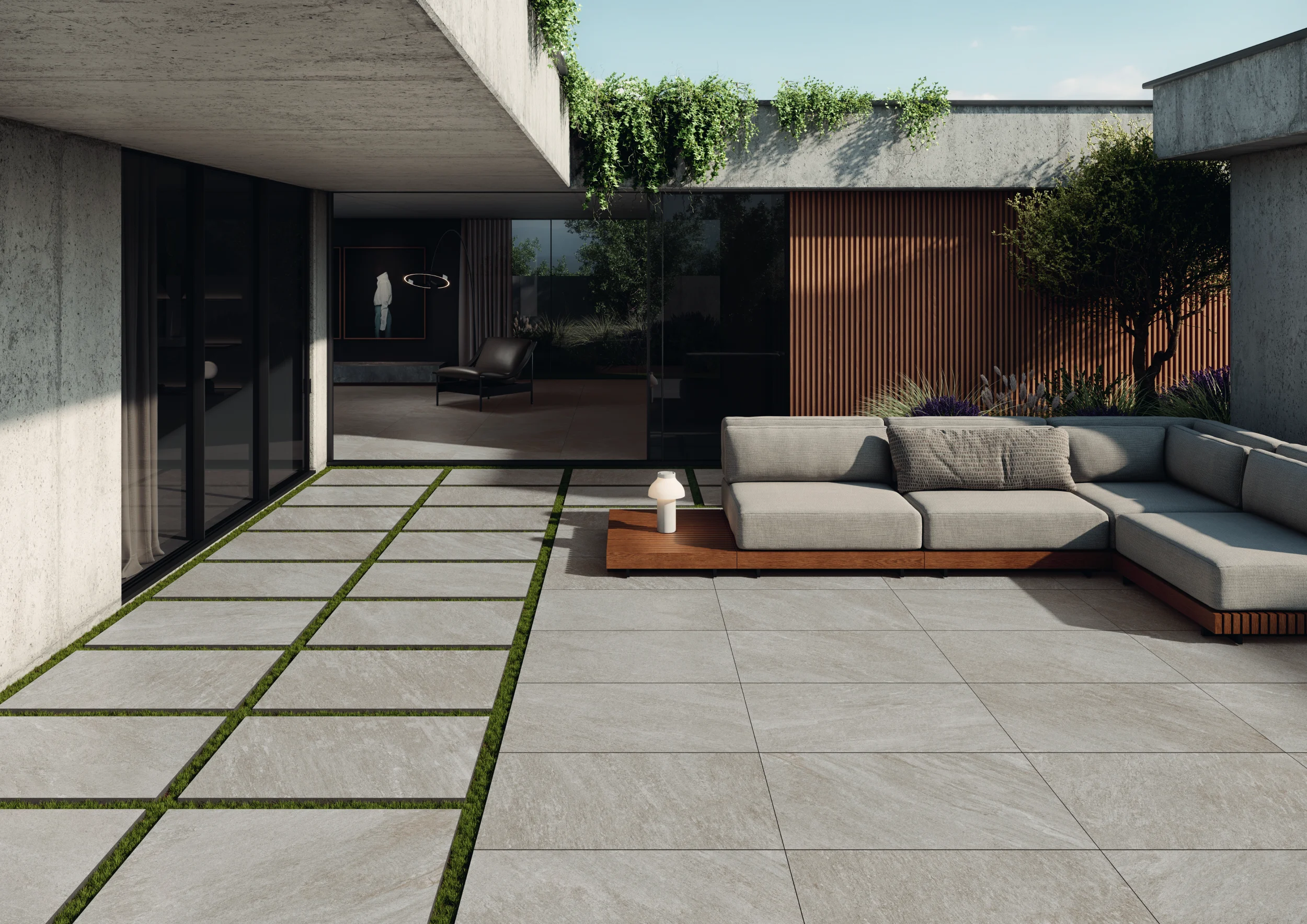 Modern outdoor patio with gray tiled floor, sectional sofa, greenery, and sliding glass doors.