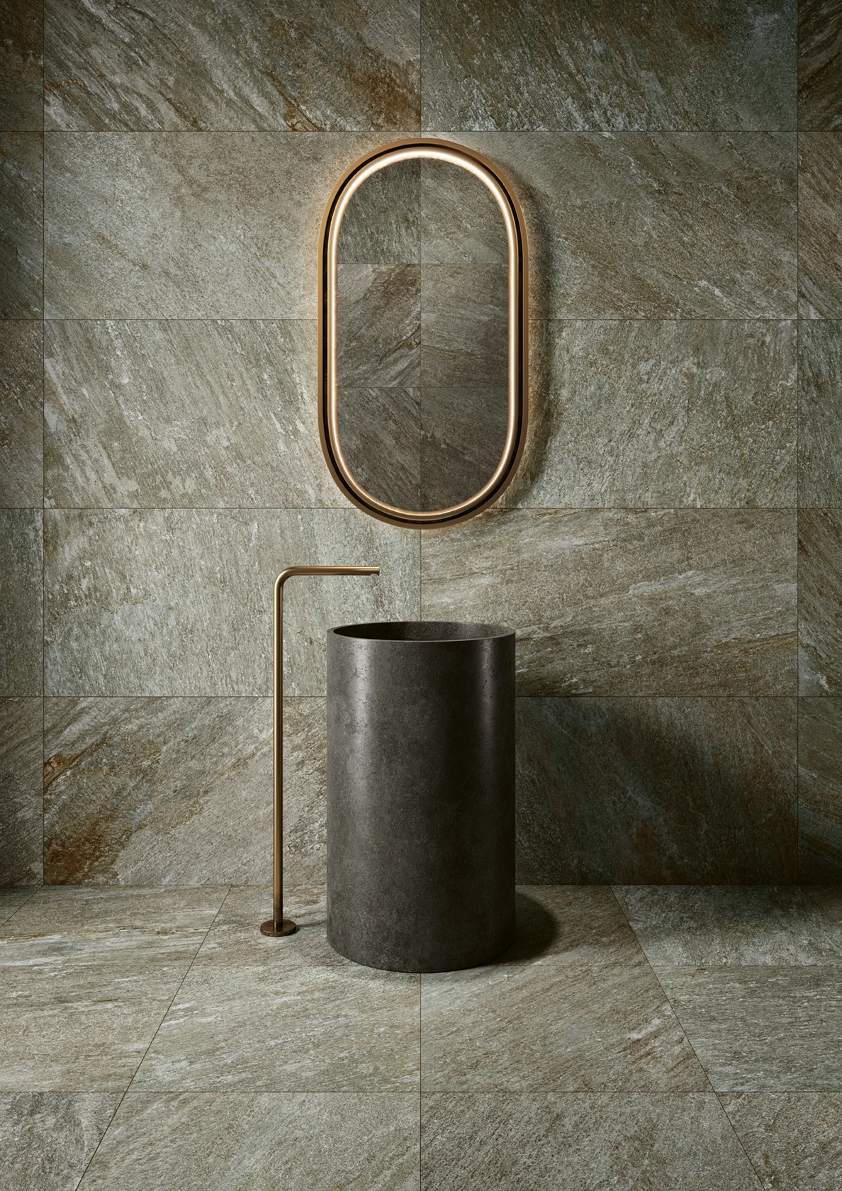 Minimalist bathroom with cylindrical dark stone sink, brass faucet, and oval brass-framed mirror on stone tile wall.