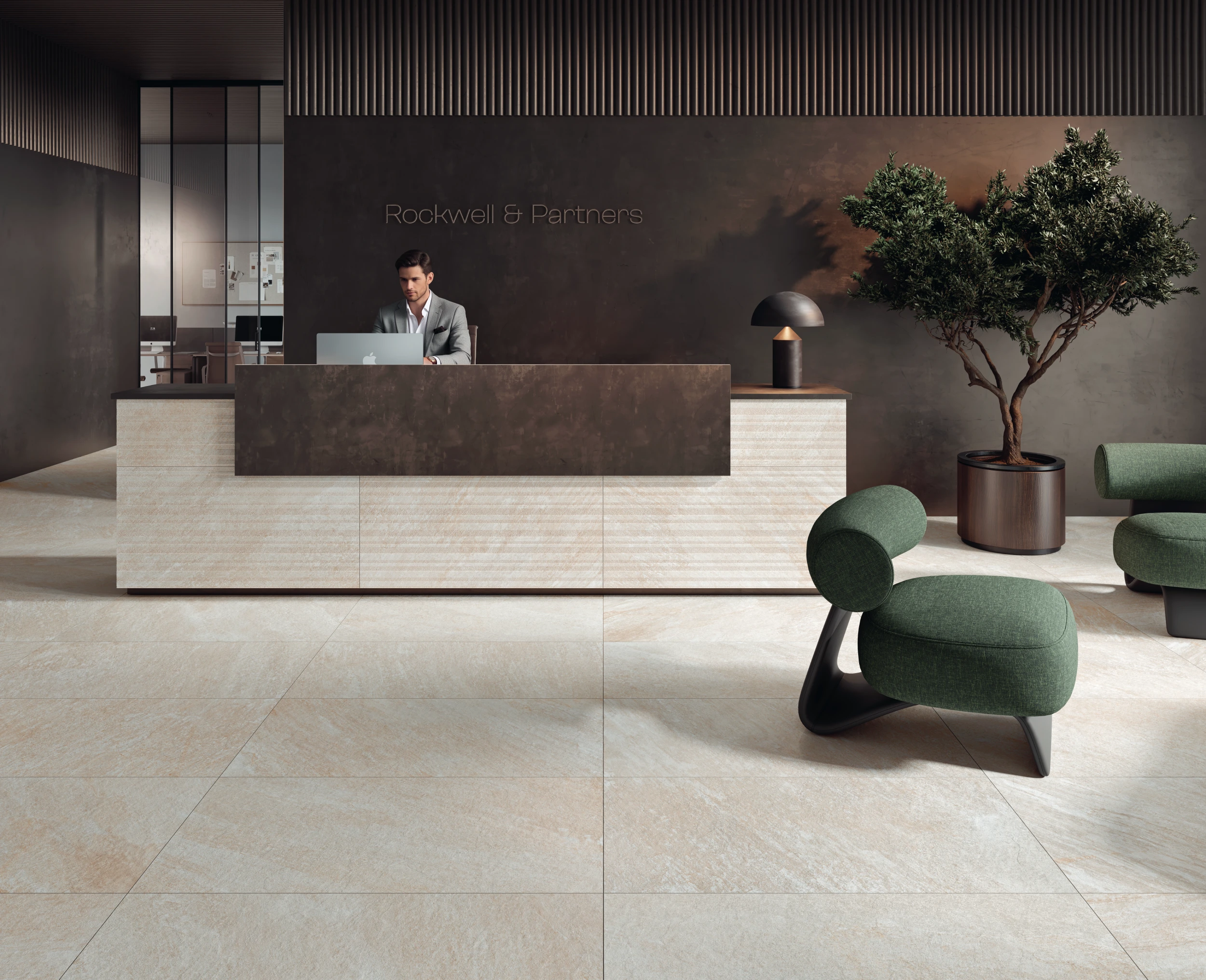 Modern office reception with a man at the desk, green chairs, a lamp, and a potted tree.