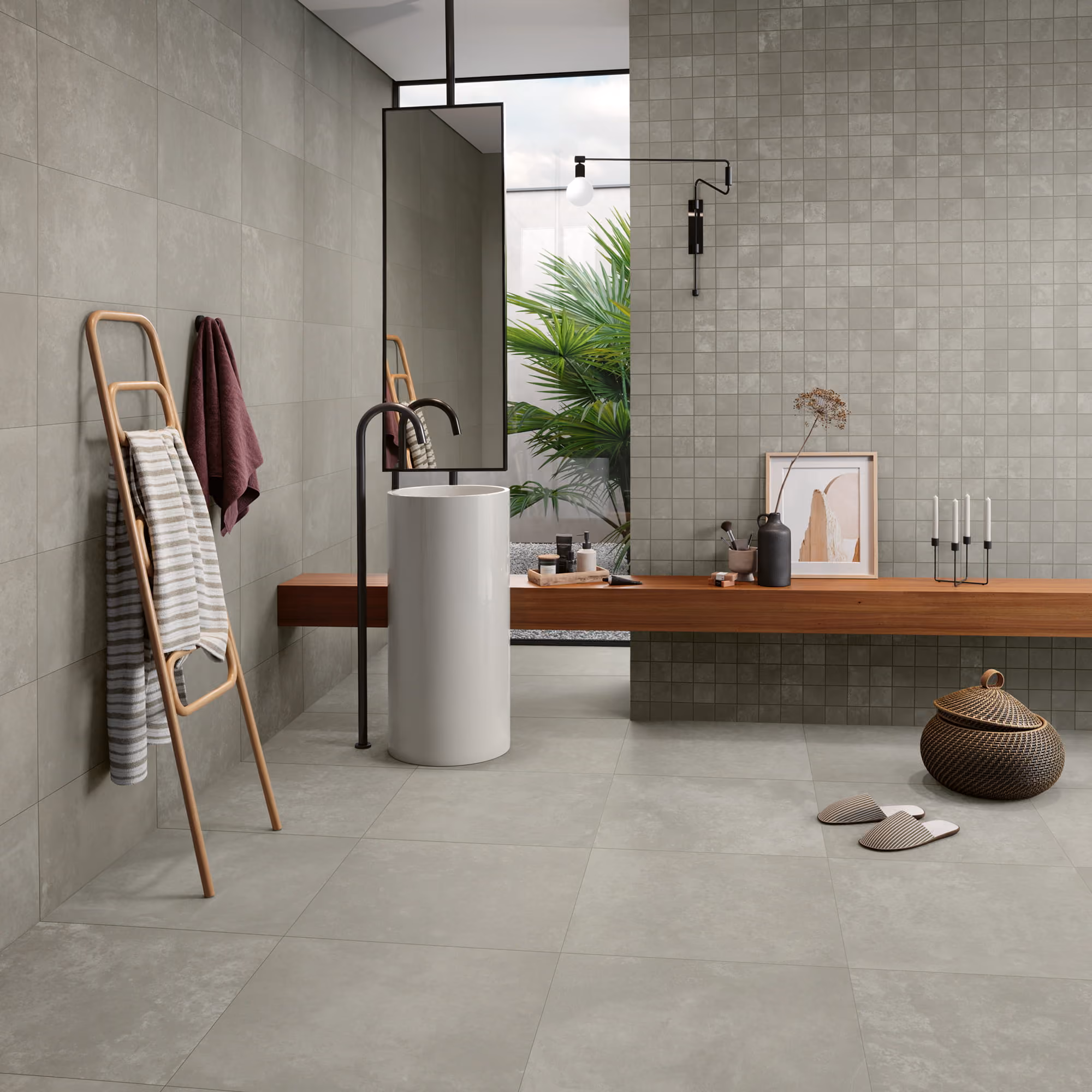 Modern bathroom with gray tiles, cylindrical white sink, wooden bench, towels, and green plant outside.