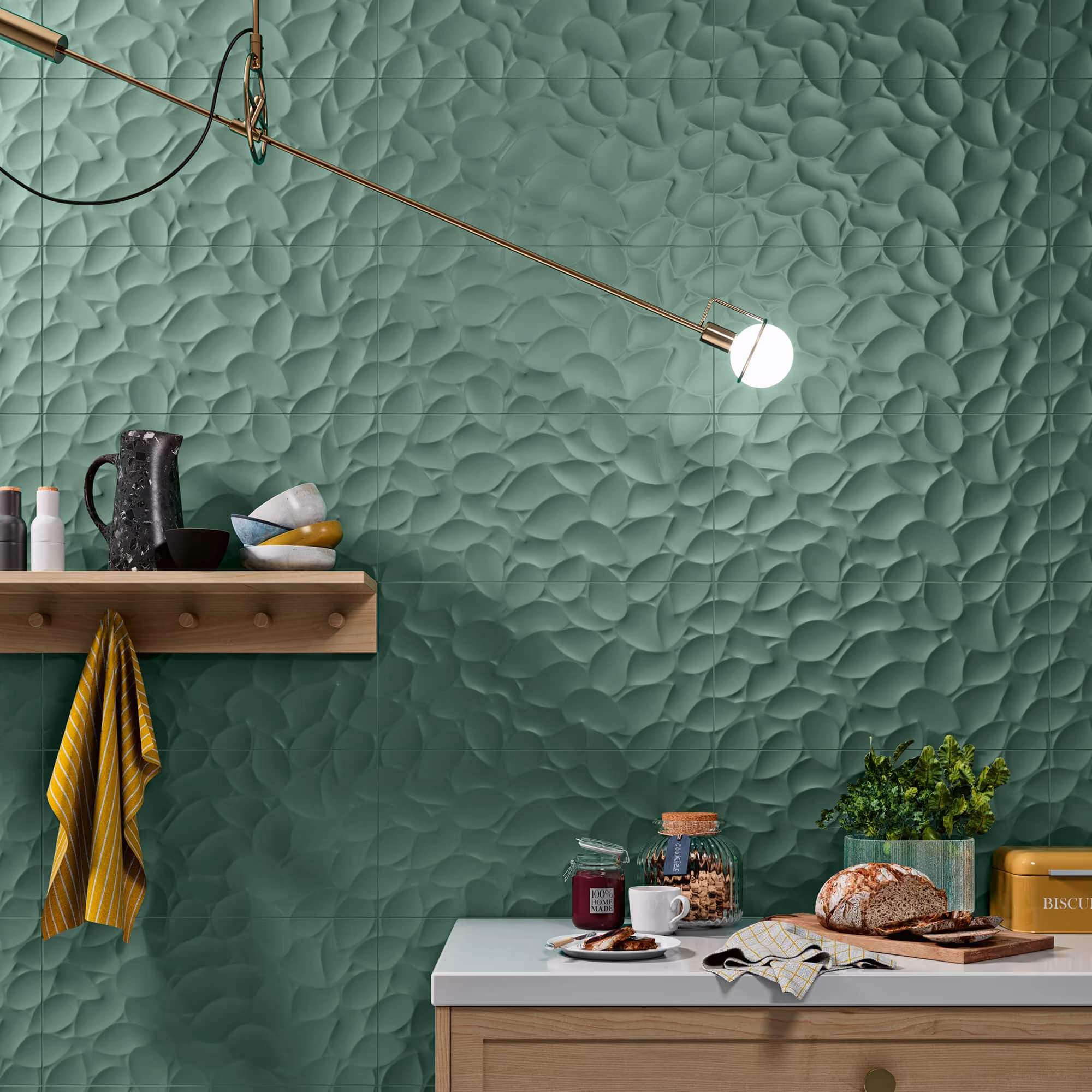 Green textured kitchen wall with wood shelf, hanging towel, and countertop with bread and plants.