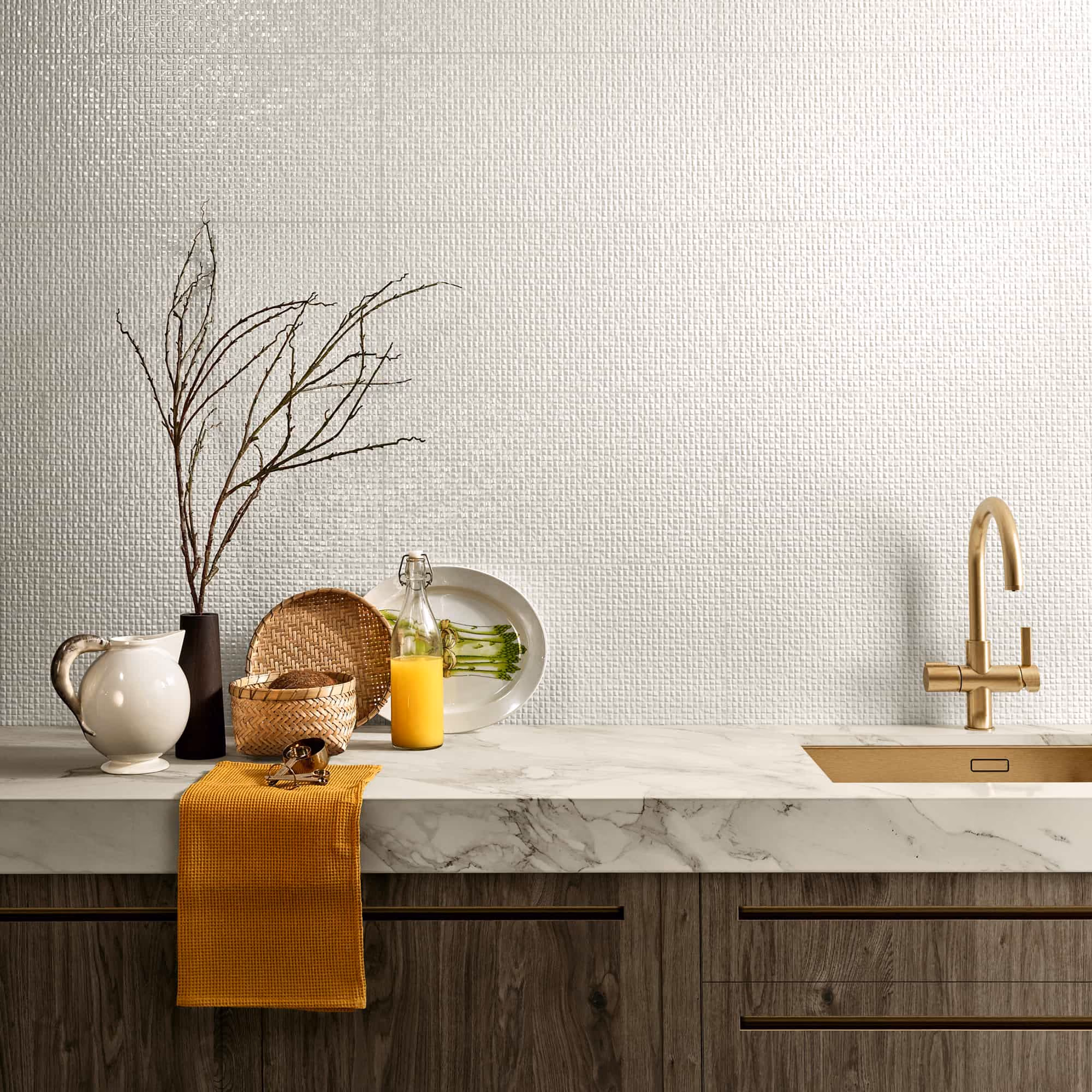 Kitchen counter with a mustard towel, vase with branches, wicker baskets, orange juice, and a gold faucet.