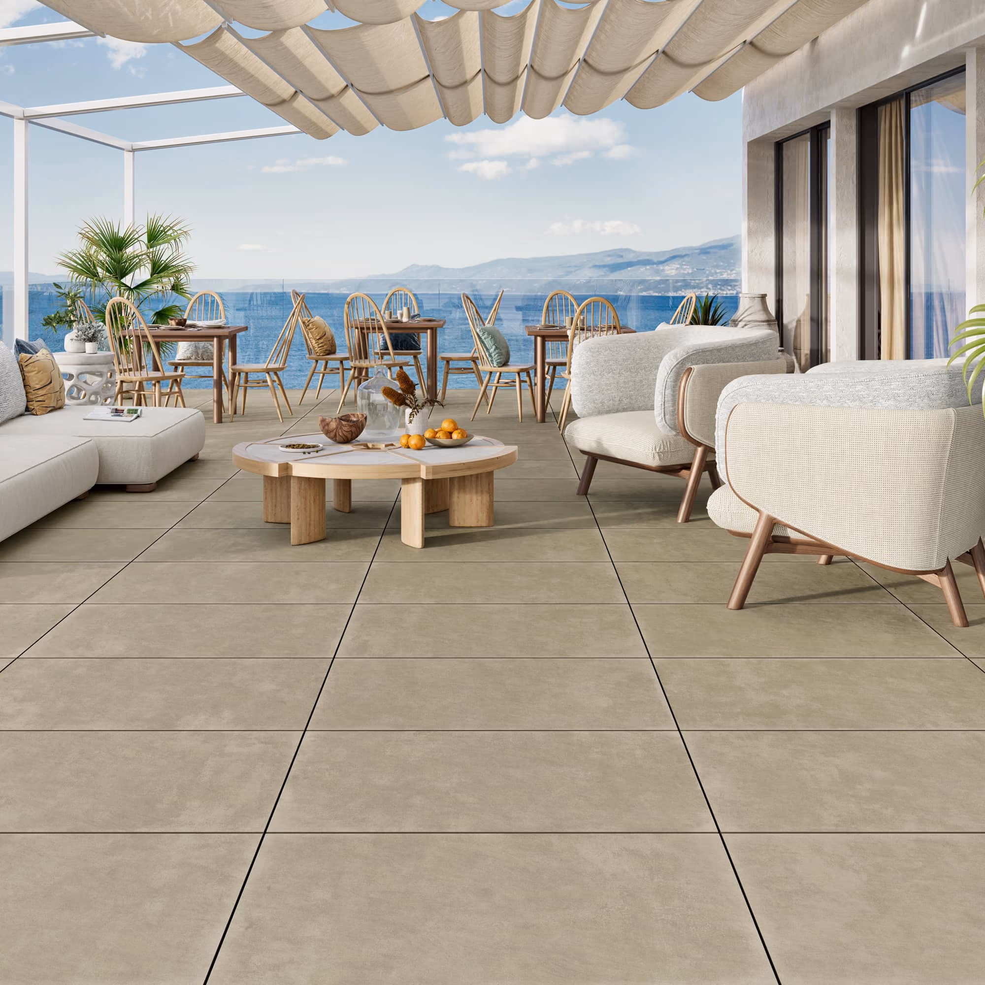 Modern outdoor patio with beige tiles, light furniture, wooden tables, and ocean view under fabric canopy.