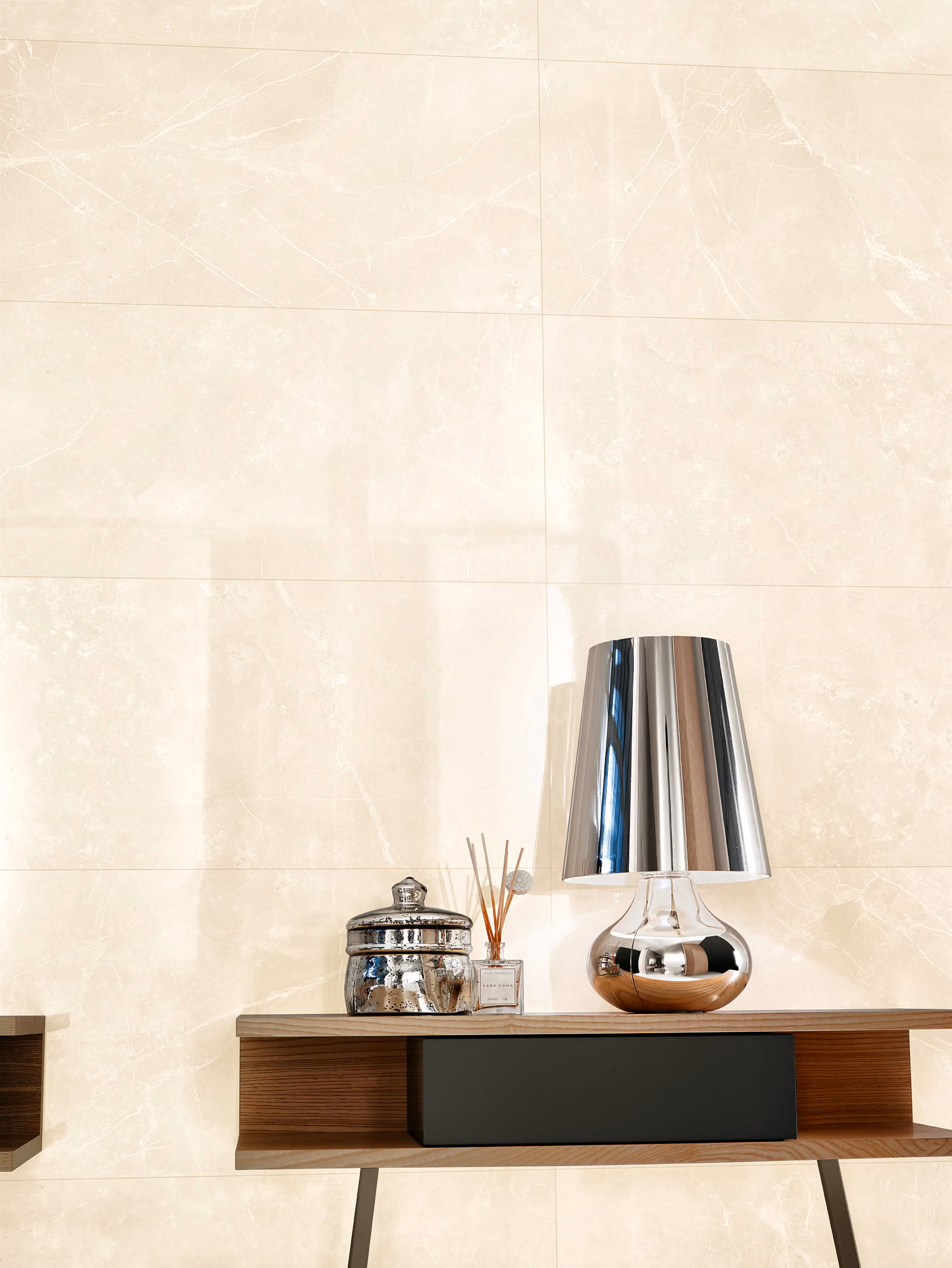 Modern wooden table with a reflective silver lamp, diffuser sticks, and a silver jar on a marble wall background.