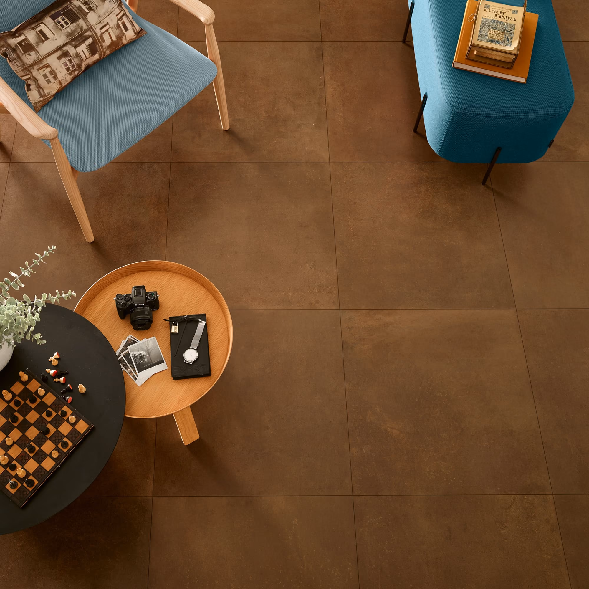 Brown tiled floor with a blue chair, teal ottoman, wooden tables, chessboard, camera, and books.