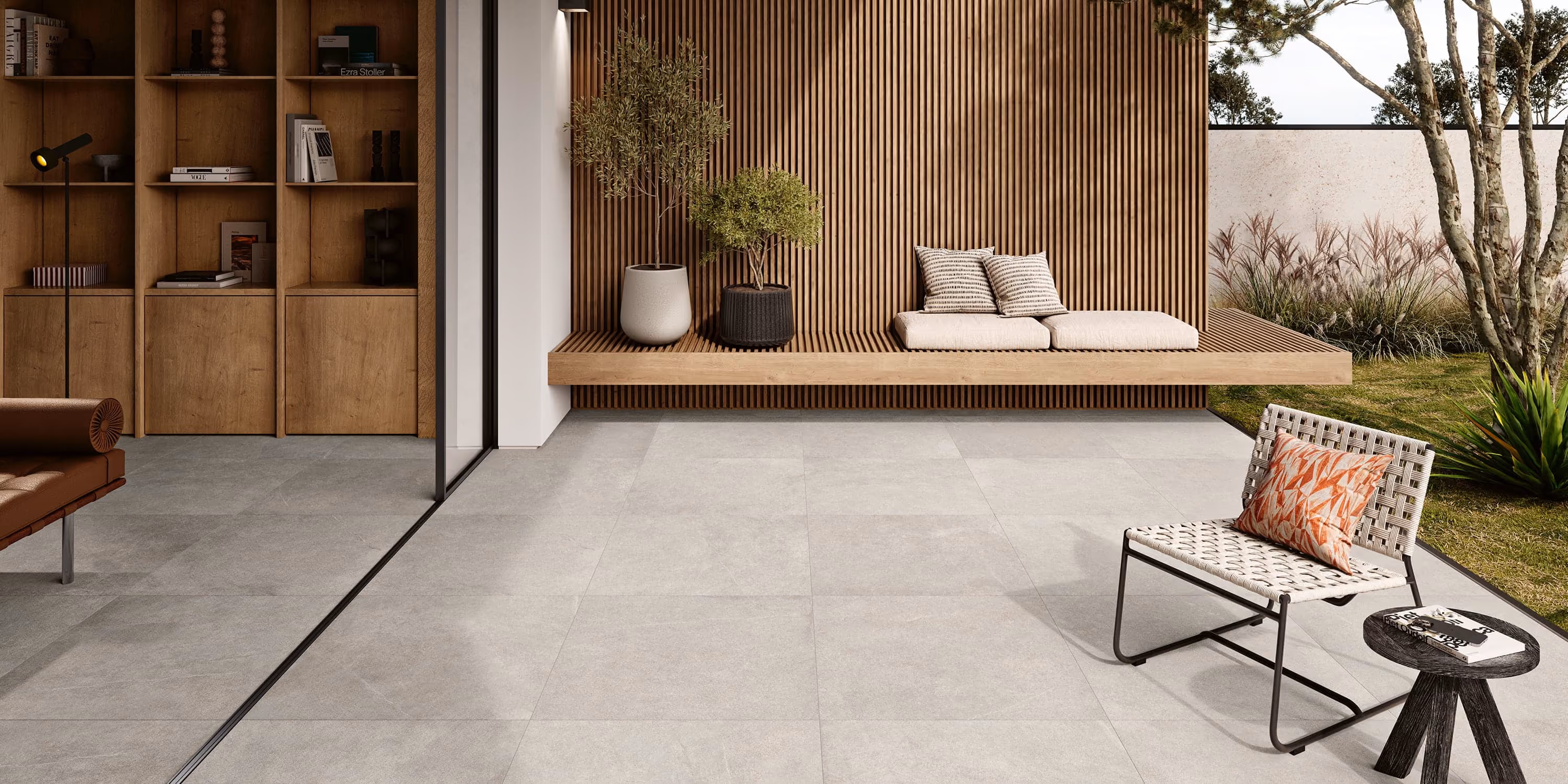 Modern outdoor patio with gray tile floor, wooden bench with cushions, potted plants, and chair with orange pillow.