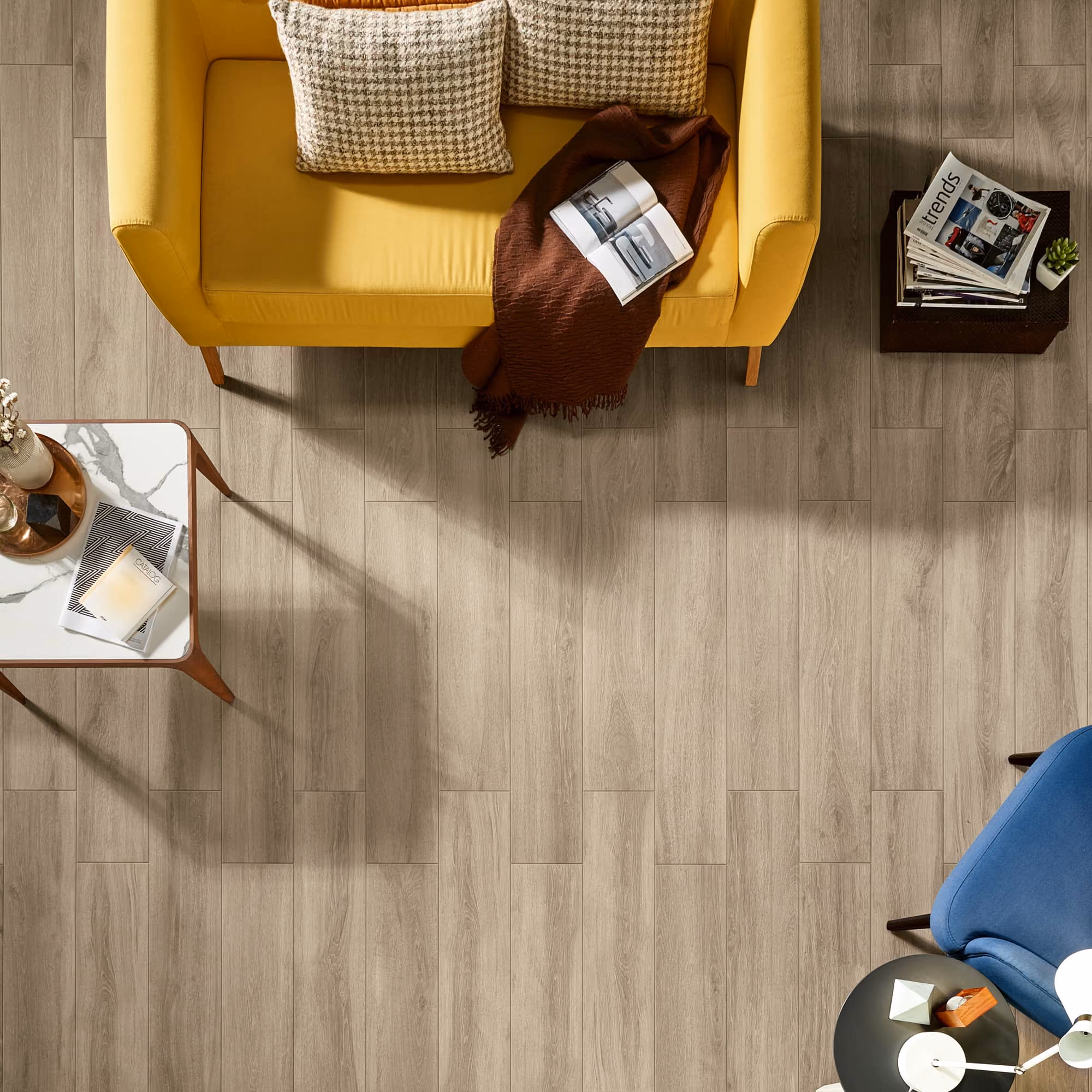 Top view of a living room with a yellow sofa, blue chair, tables, and light wood floor tiles.