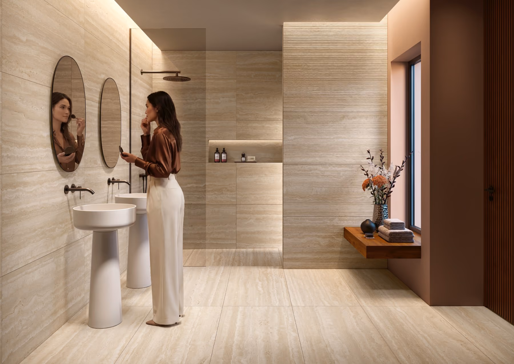 Woman applying makeup in a modern bathroom with beige tiles, dual sinks, and a flower vase by a window.