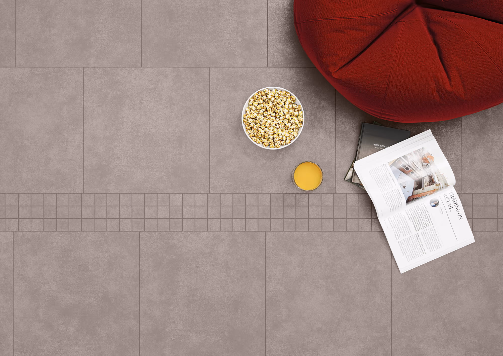 Top view of a bowl of popcorn, glass of orange juice, open magazine, and red bean bag on tiled floor.