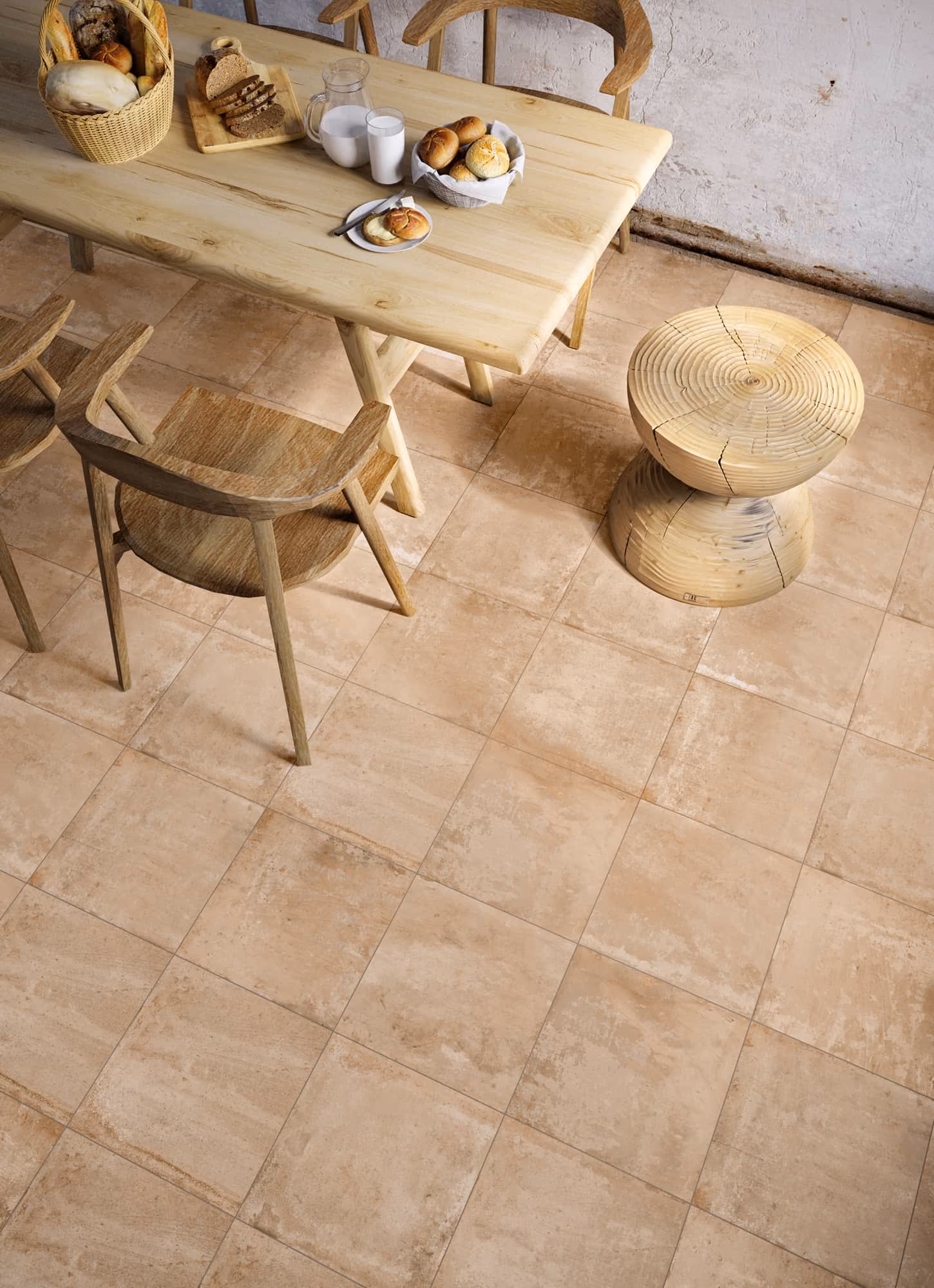 Dining table with bread, milk jug, basket, wooden chairs, and a wooden stool on tiled floor.
