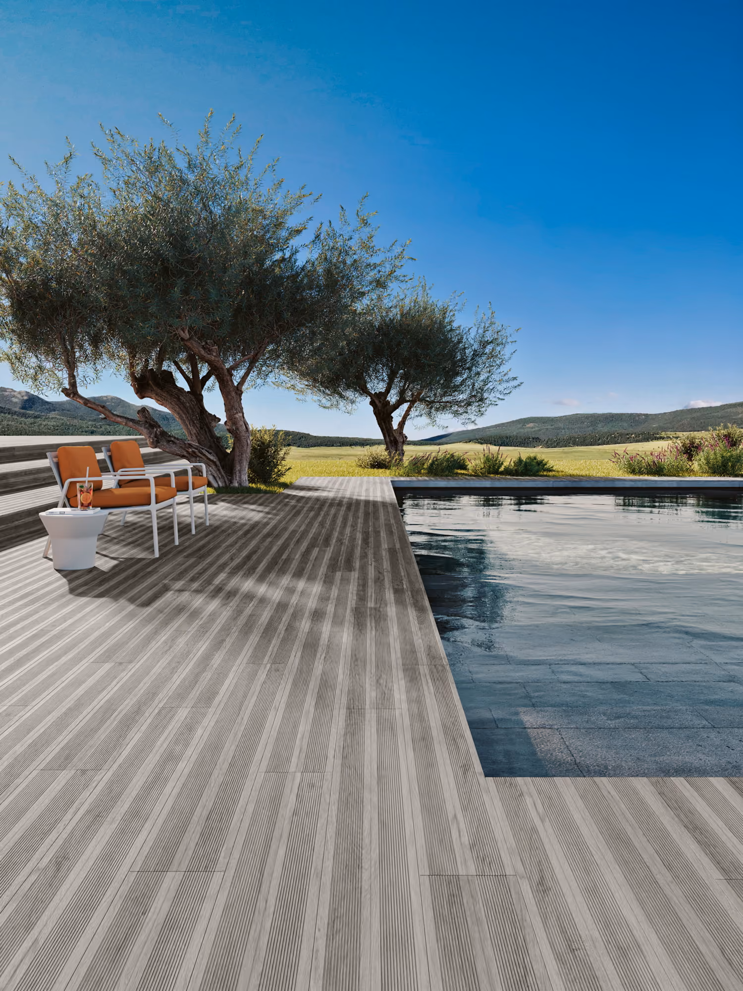 Wooden deck with two chairs and a drink next to a pool and olive trees under a clear blue sky.