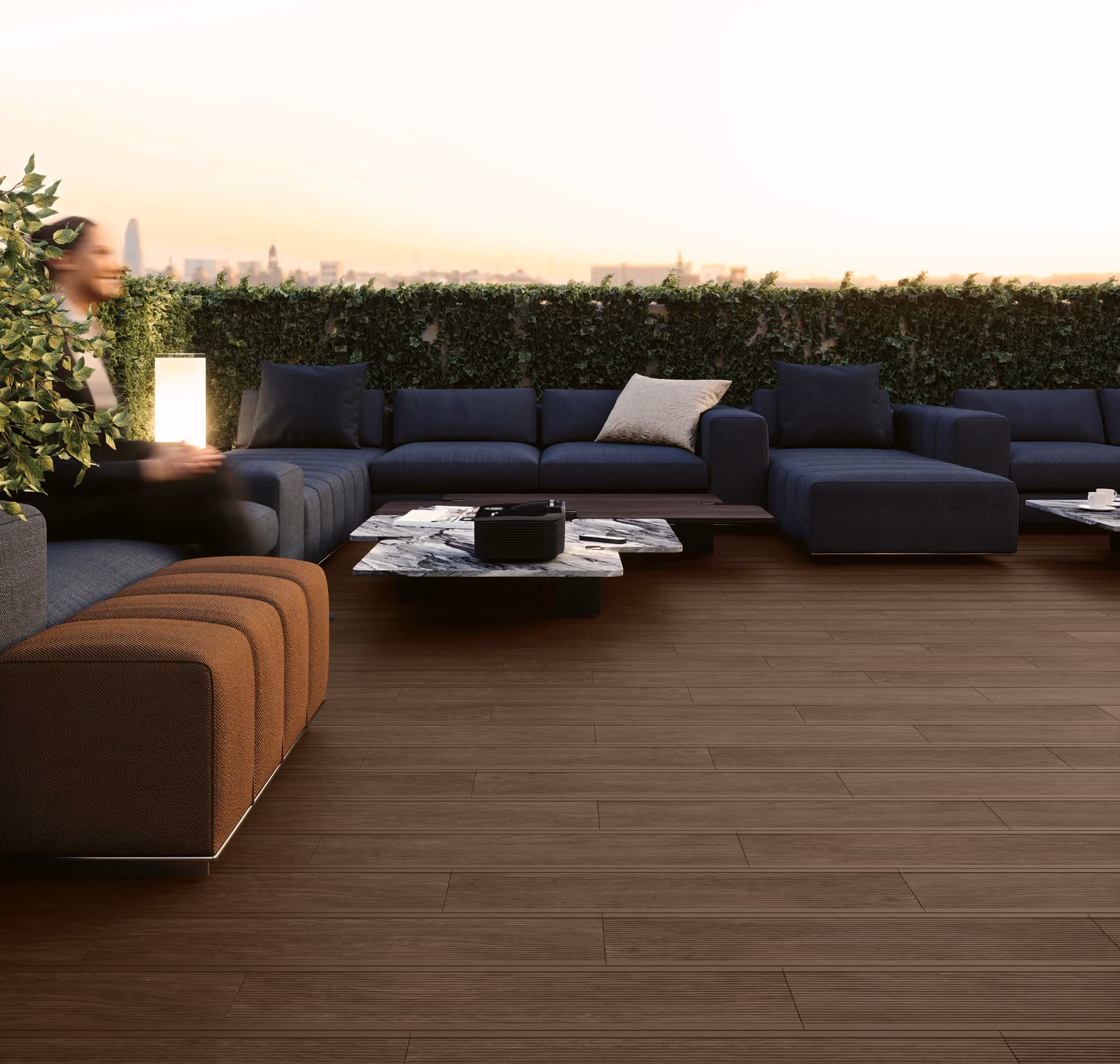 Modern outdoor seating area with dark sofas, marble tables, a glowing lamp, and a hedge wall at sunset.