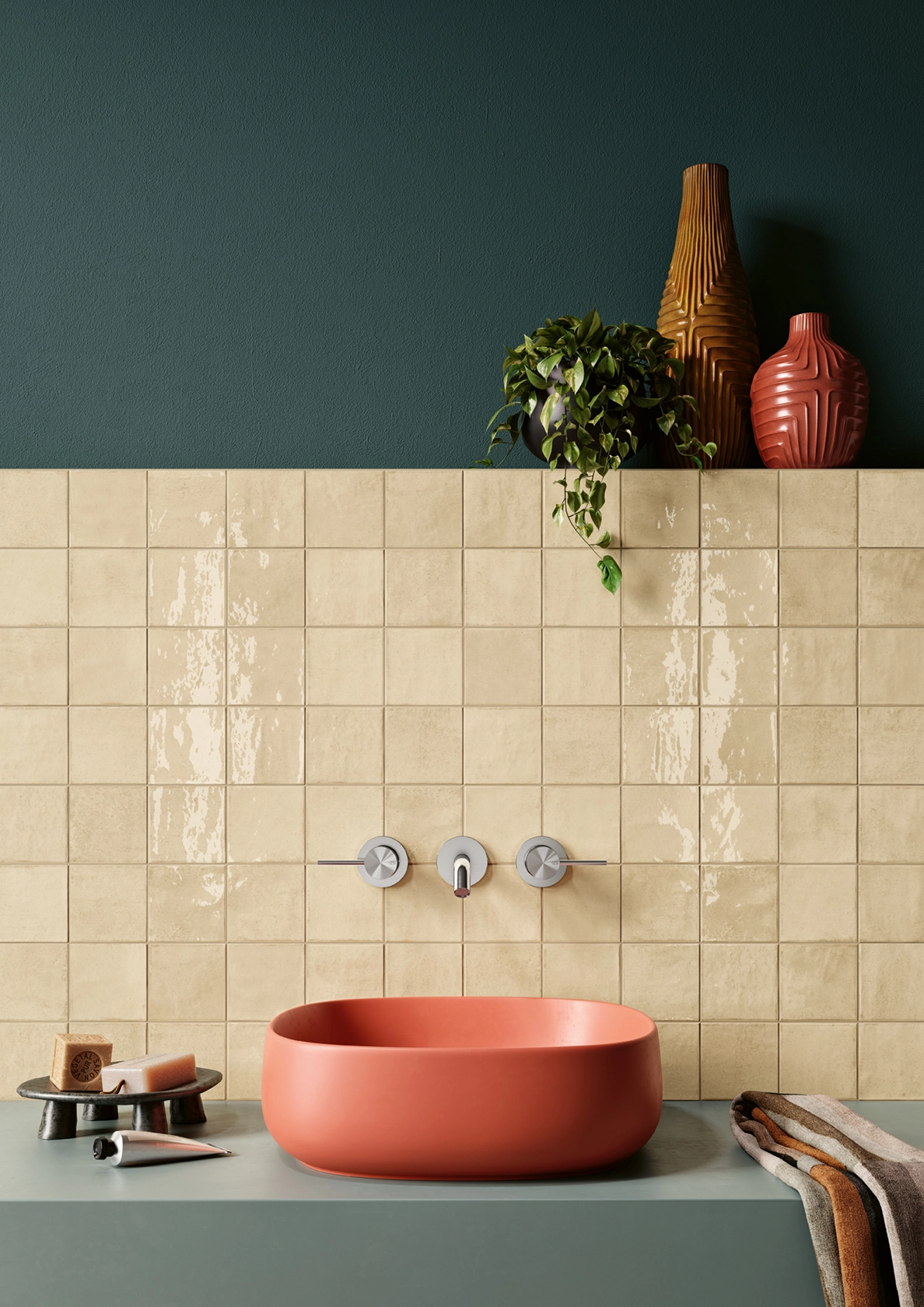 Orange oval sink with chrome faucet set in a green countertop, beige tiled wall, plants, and towels.