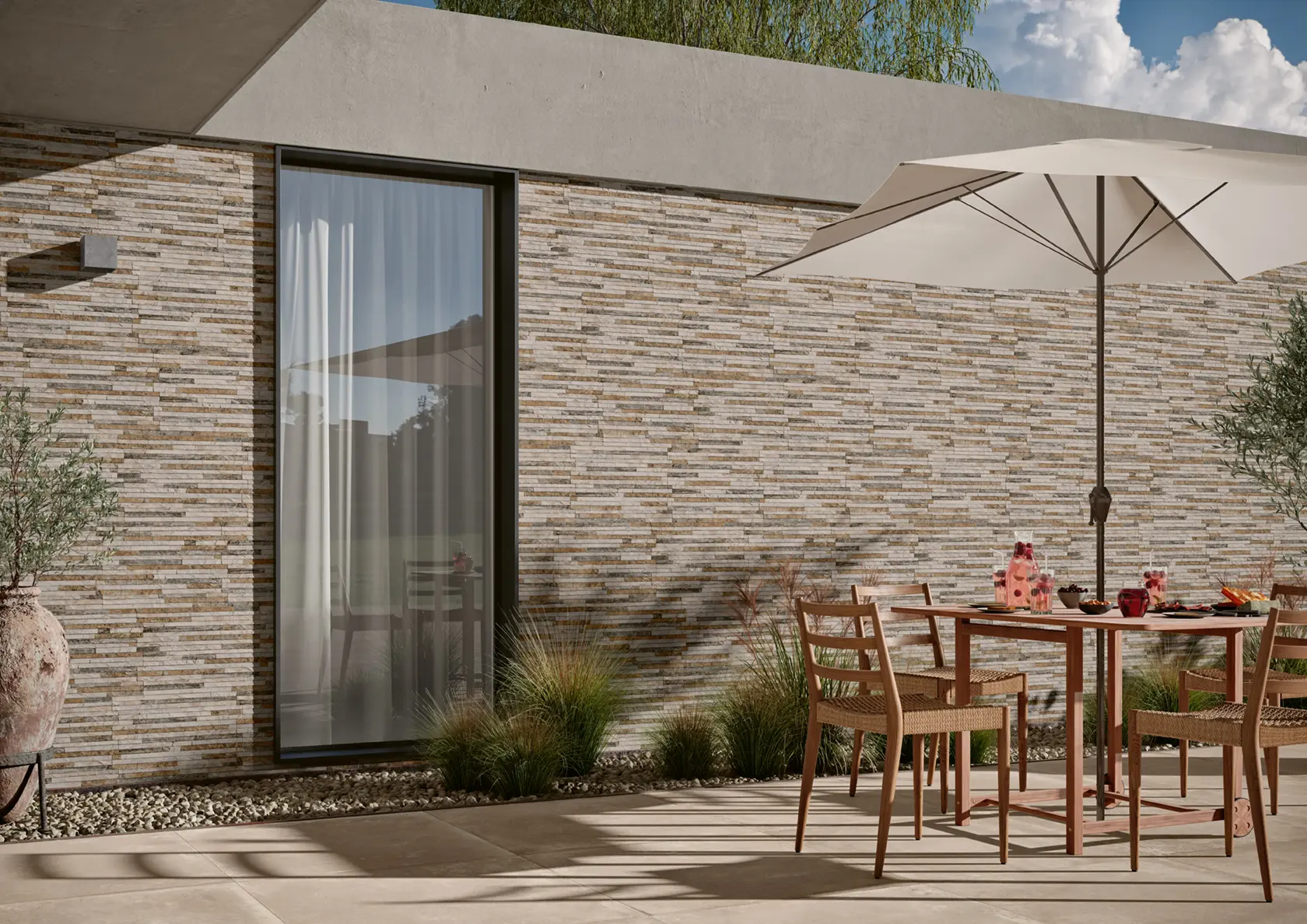 Outdoor patio with a wooden table, four chairs, umbrella, and drinks against a stone wall.