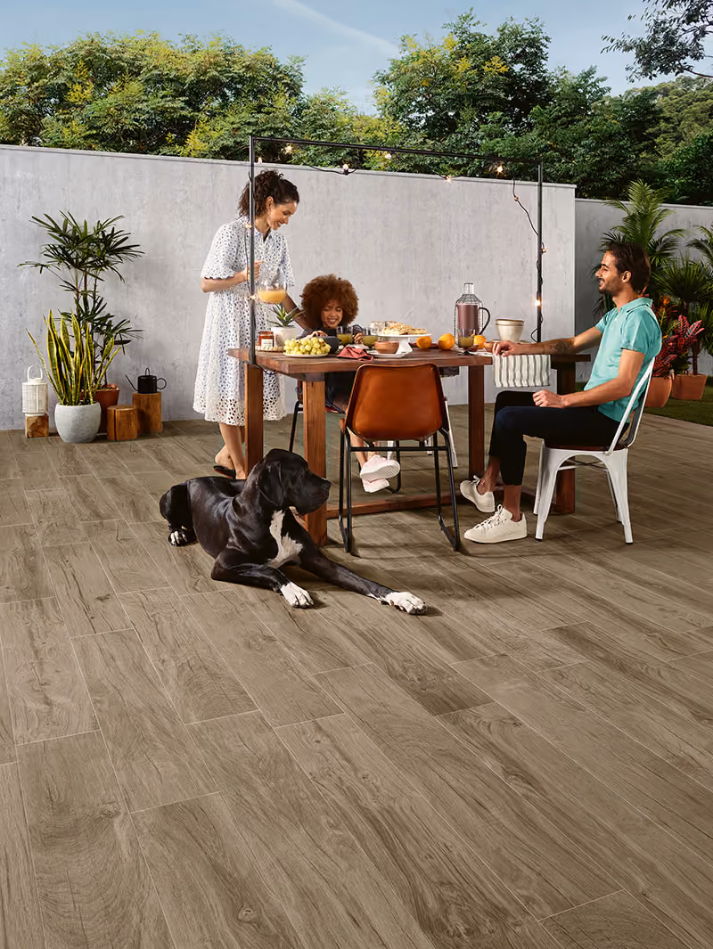 Three people and a black dog at an outdoor table with food and plants around on wood floor.