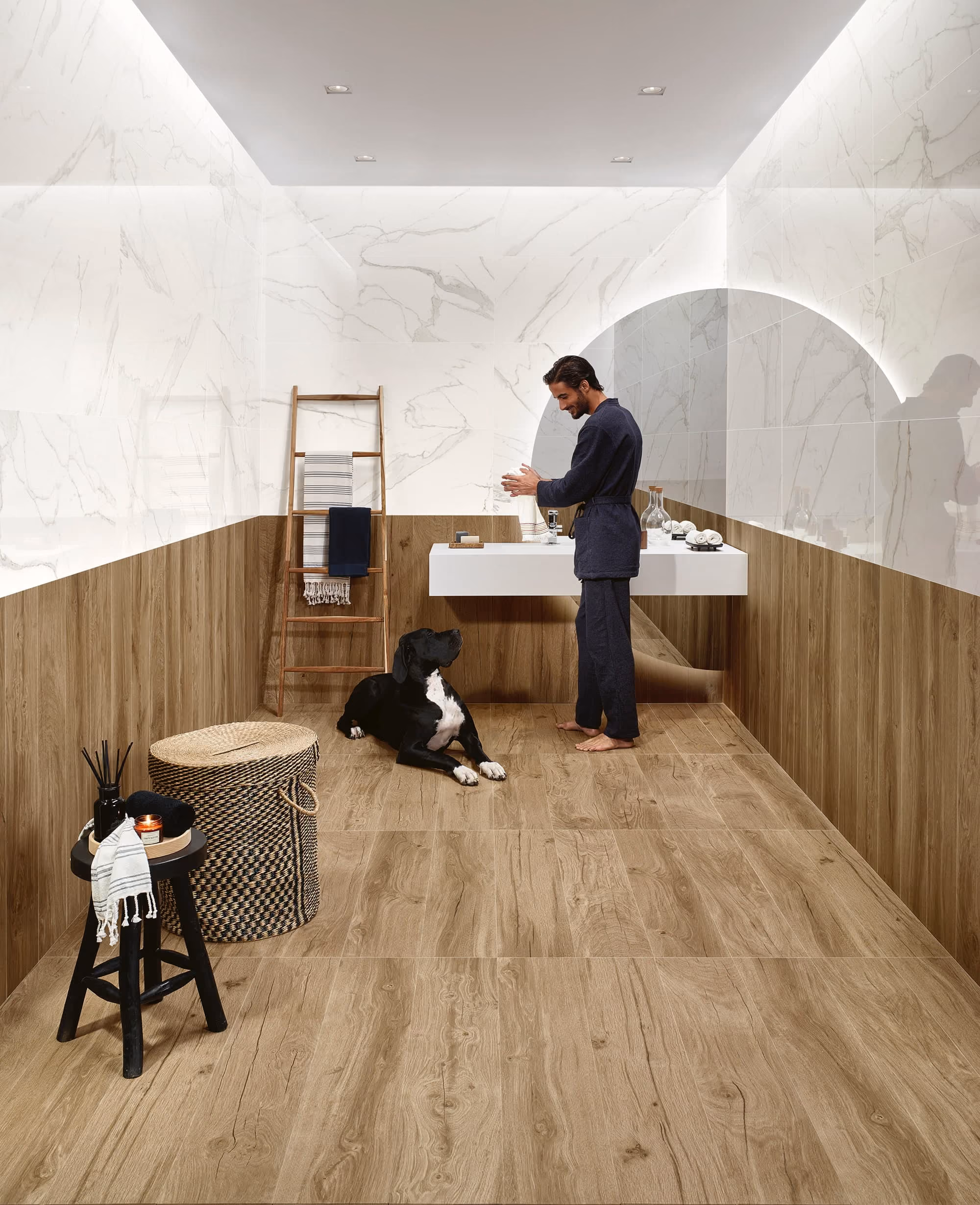 Man in dark robe stands barefoot in a modern bathroom with wood and marble tiles and a black dog.