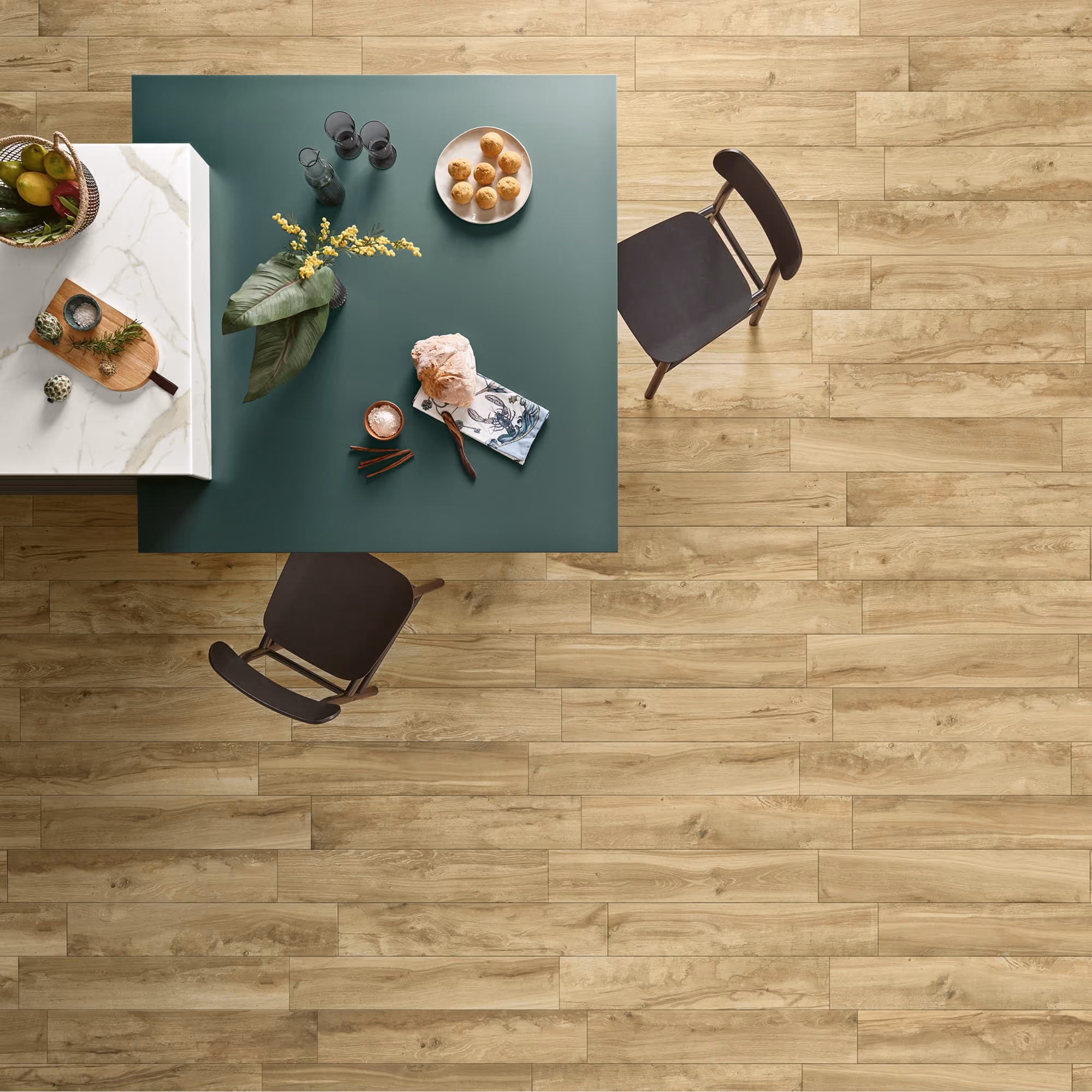 Top view of green table with bread, flowers, glasses, and two chairs on light wood floor.