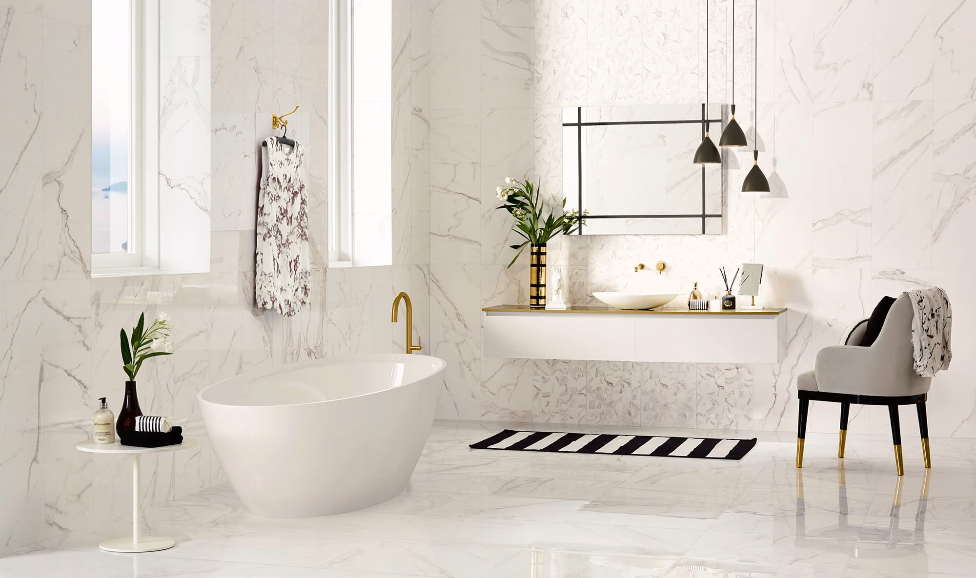 Modern white marble bathroom with freestanding tub, gold fixtures, chair, and black-and-white striped rug.