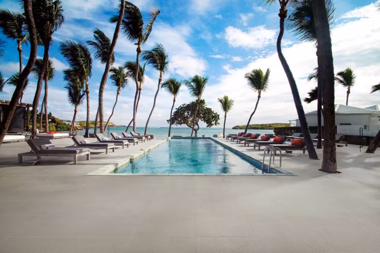 Outdoor pool with lounge chairs and palm trees beside a beach under a partly cloudy sky.