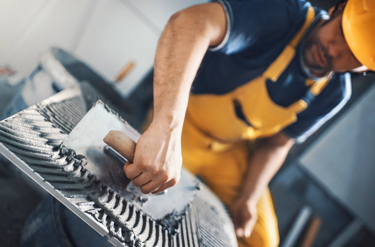 Construction worker in yellow overalls spreading cement with a trowel on a ribbed surface.