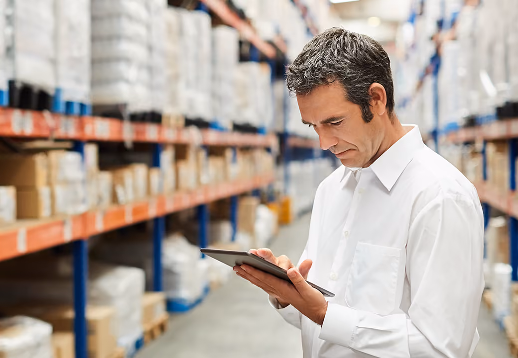 Man in white shirt using tablet in a warehouse with shelves of boxed goods.