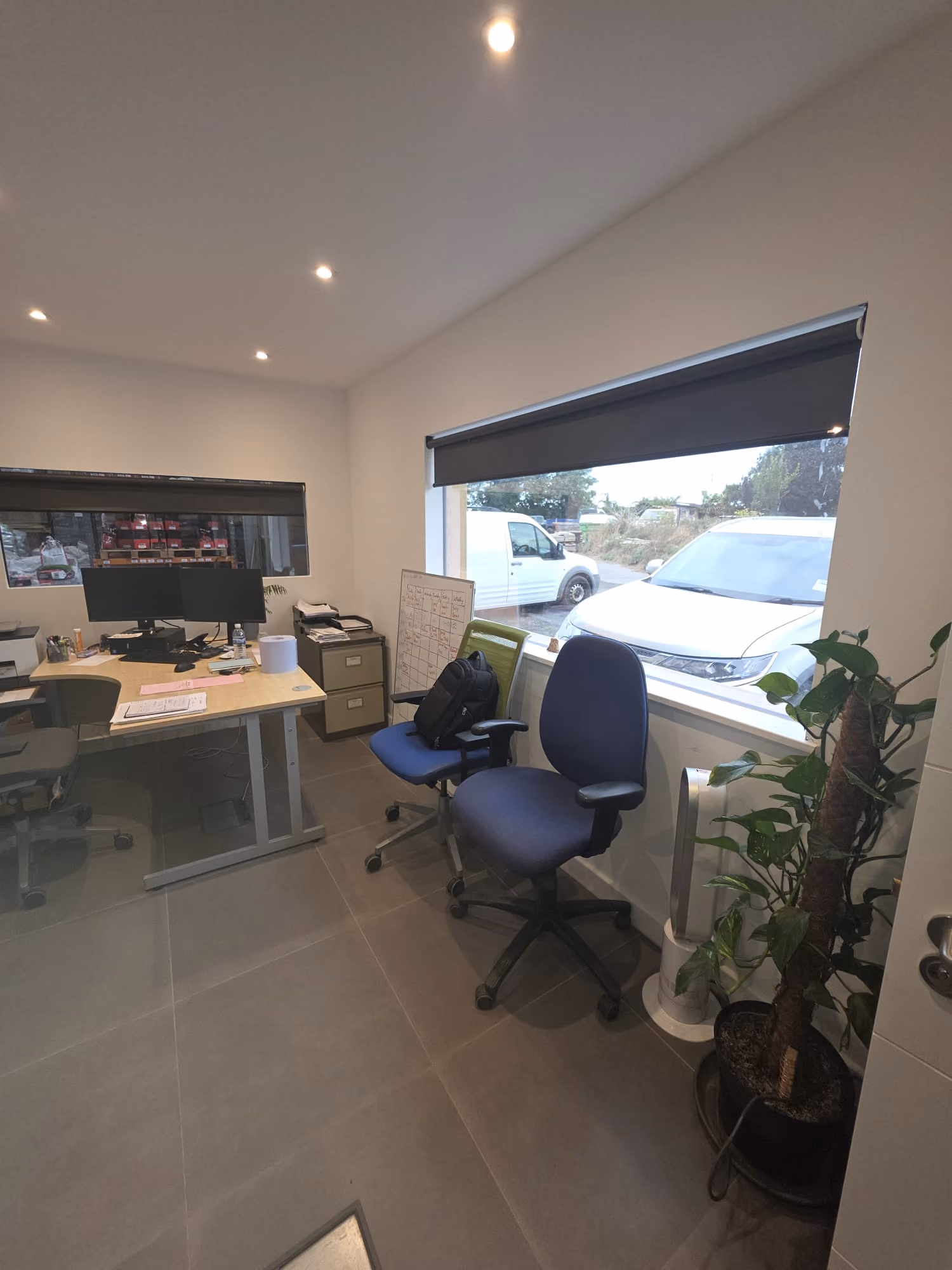 Small office with desk, computers, chairs, filing cabinet, and window showing parked vehicles outside.
