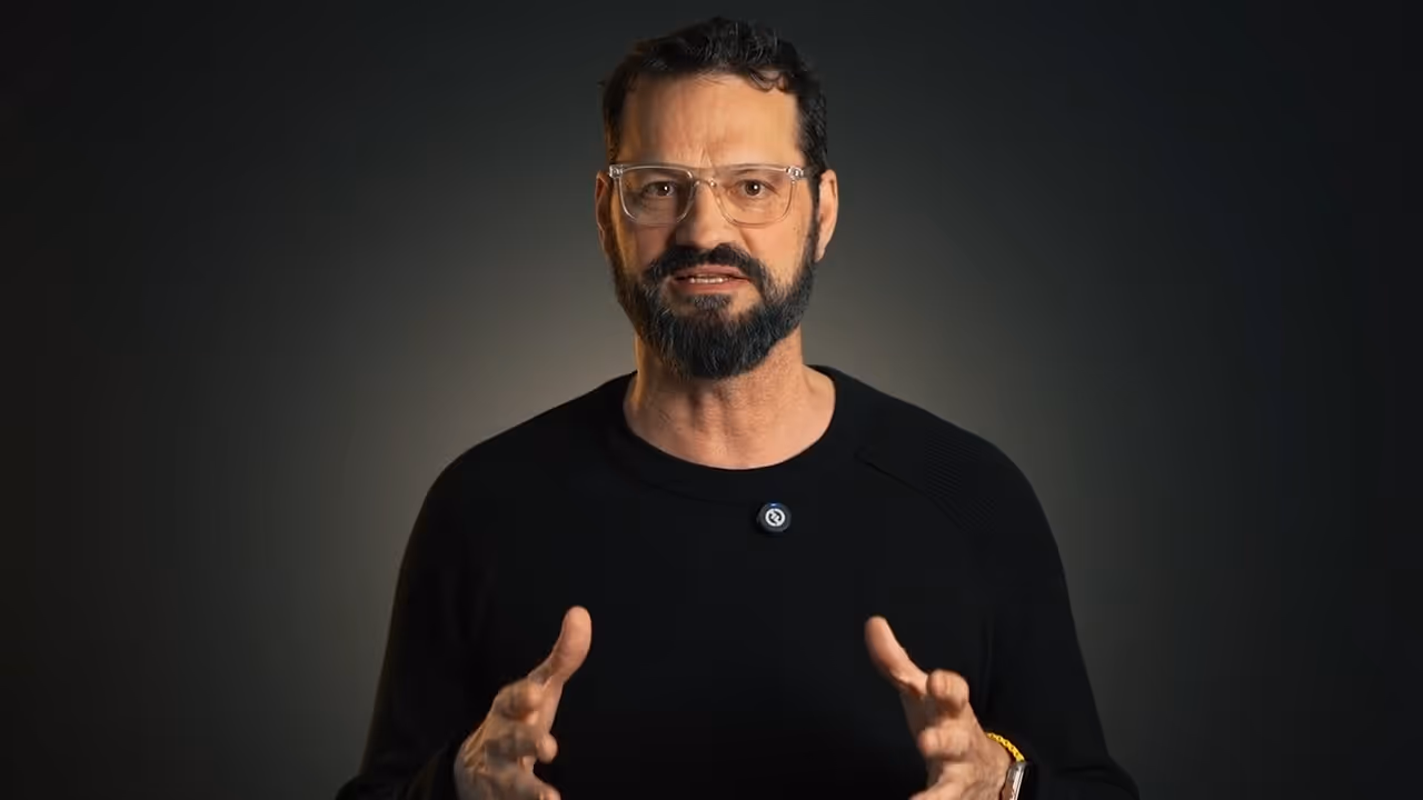 Man with glasses and beard speaking with hands raised against a dark gradient background.