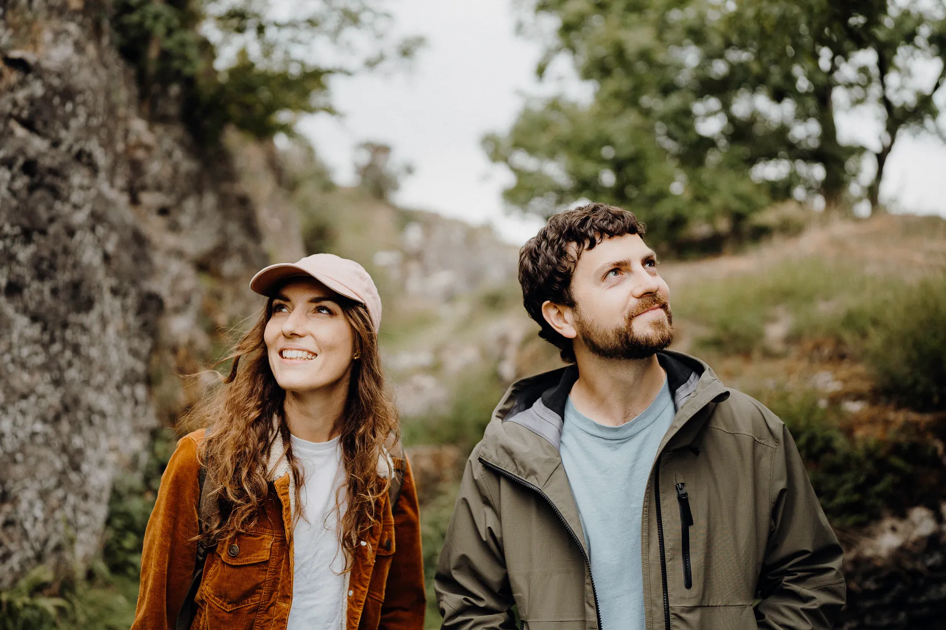 Smiling couple outdoors in jackets looking up amid rocky terrain and trees in background.