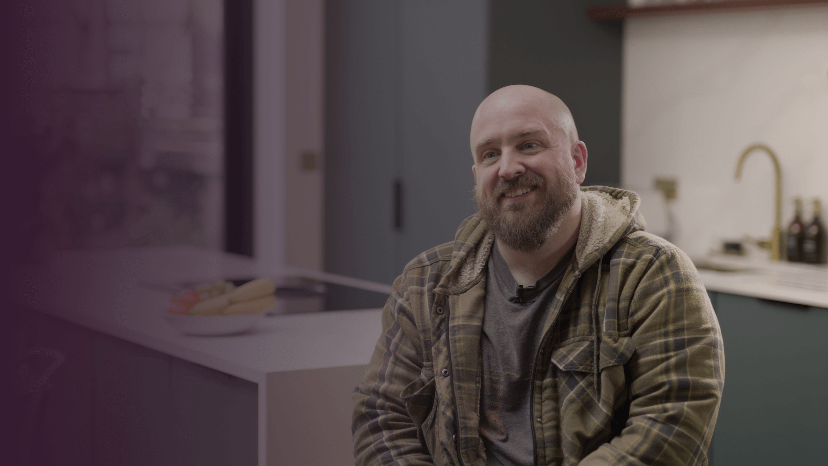 Smiling man with a beard wearing a flannel jacket, sitting in a modern kitchen.