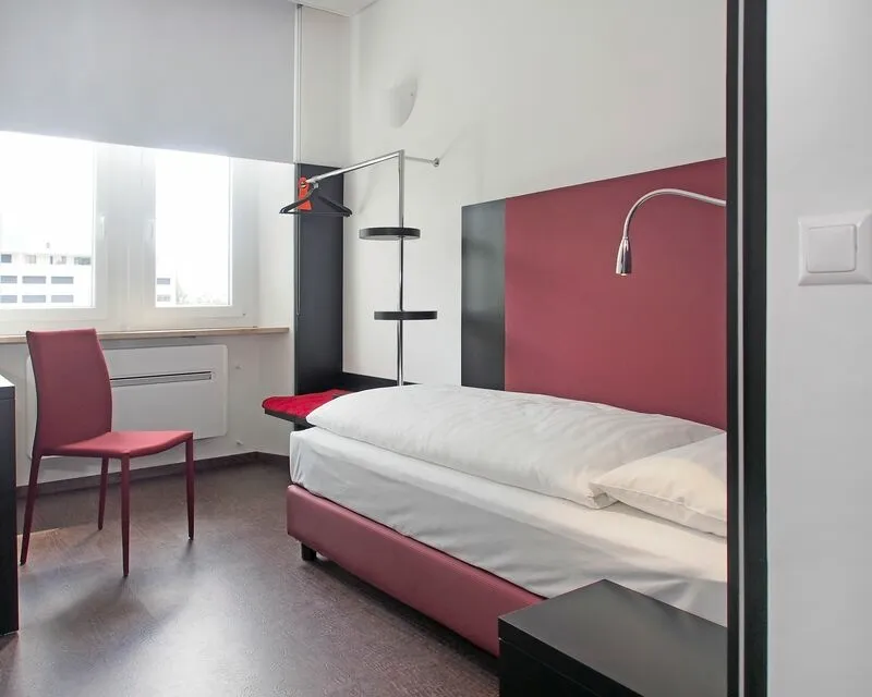 Strakke hotelkamer met eenpersoonsbed, rode accenten, bureau en stoel bij het raam