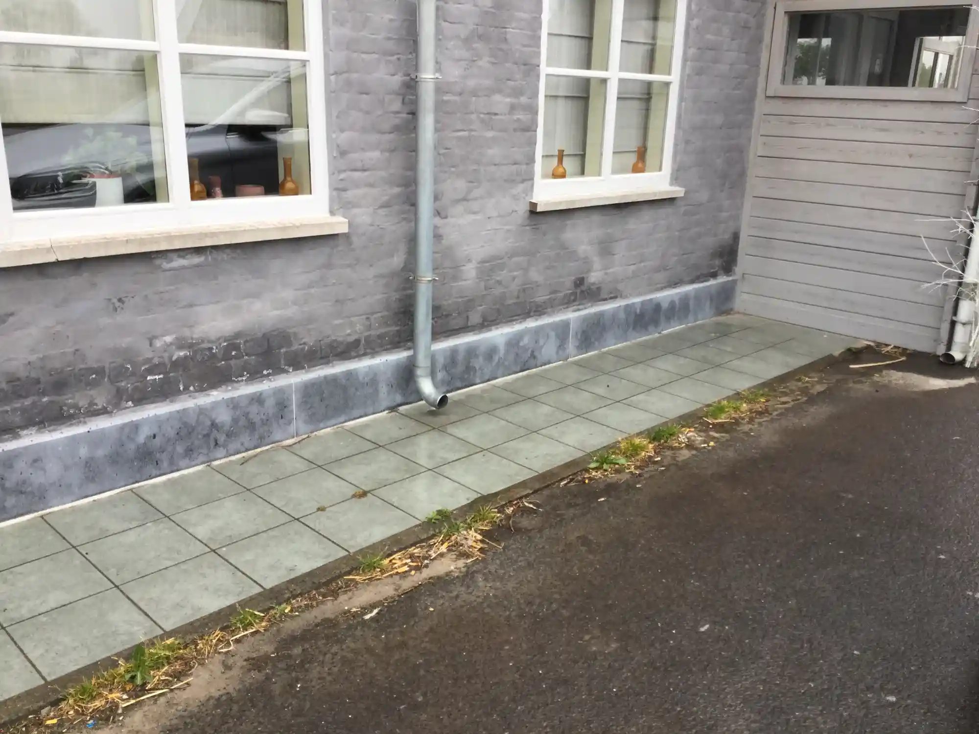 Grijze gevelvoet met regenpijp en nieuwe zinken plint langs een betegeld pad