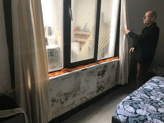 Man kijkt naar een raam met condens en zwarte schimmel op de muur eronder