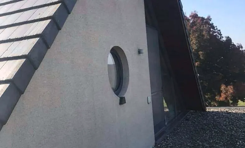 Moderne gevel met grijze crepi-afwerking en rond raam, naast een hellend dak met leien
