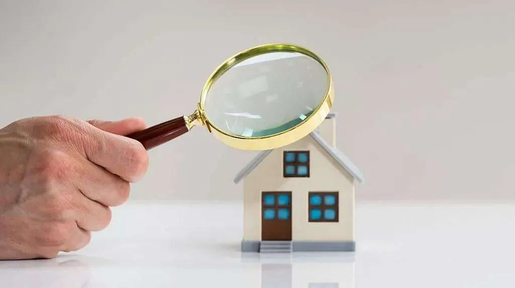 Hand met vergrootglas onderzoekt een miniatuurhuisje, symbool voor woninginspectie