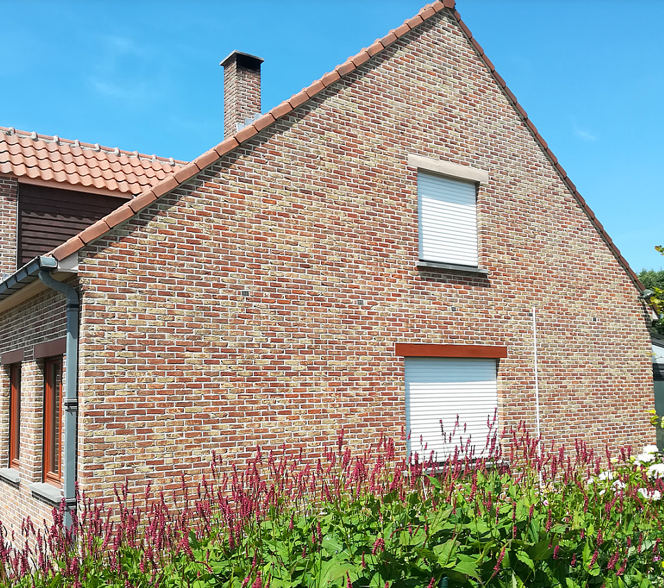 Schone bakstenen zijgevel van een woning na gevelreiniging, met witte rolluiken en bloeiende planten
