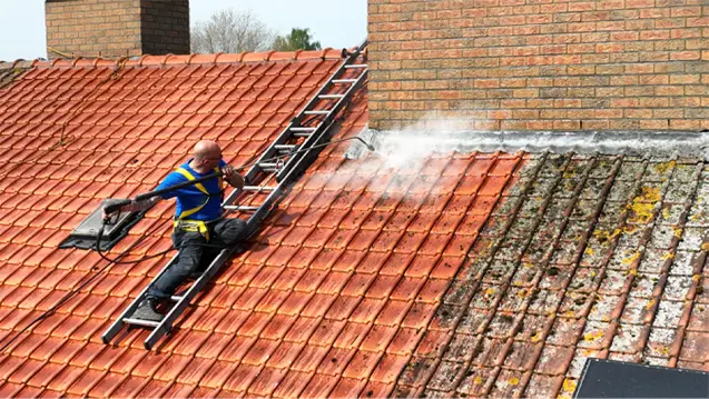 Vakman met harnas op een ladder reinigt een bemost pannendak met een hogedrukreiniger