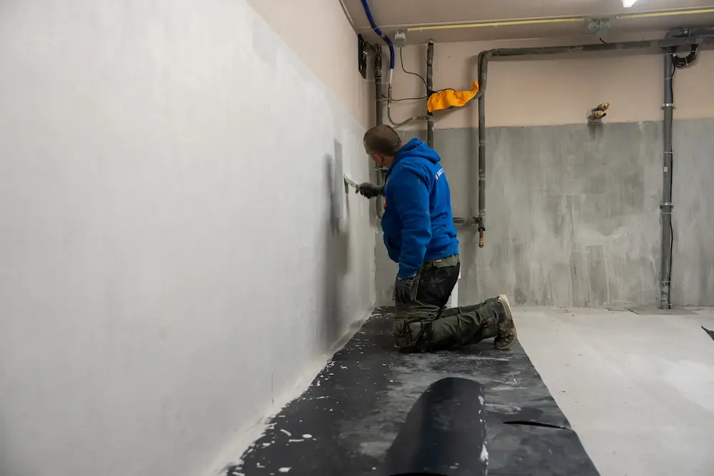 Vakman werkt waterdichte coating af op een keldermuur, met dichtingsmembraan op de vloer