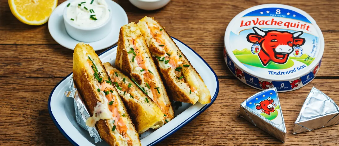 Trois sandwichs grillés au fromage fondu et saumon, garnis de ciboulette, servis dans un plat blanc à bord bleu, avec un demi citron, sauce blanche, et fromage La Vache qui rit à côté.