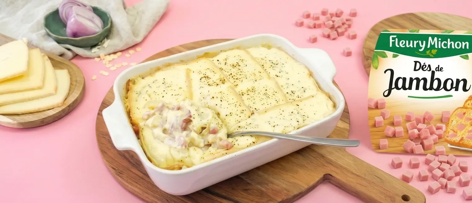 Gratin au jambon avec tranches de fromage fondu dans un plat blanc sur une planche en bois, accompagné de dés de jambon Fleury Michon et d'oignons.