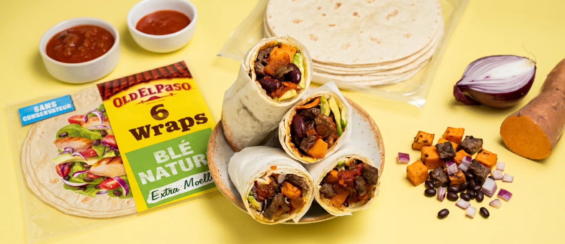 Trois wraps garnis de légumes, viande et haricots dans un bol à côté d'un paquet de wraps Old El Paso, sauces, oignon rouge, patate douce et haricots noirs sur fond jaune.