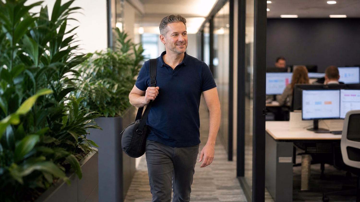 Man in navy polo walks down plant-lined office corridor
