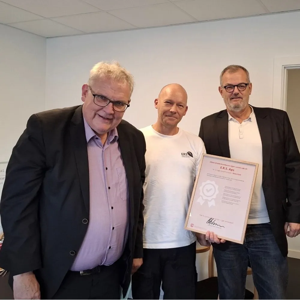 Tre mænd, hvor en holder et indrammet Diplom fra Albertslund Kommune til E.R.S i et kontormiljø.