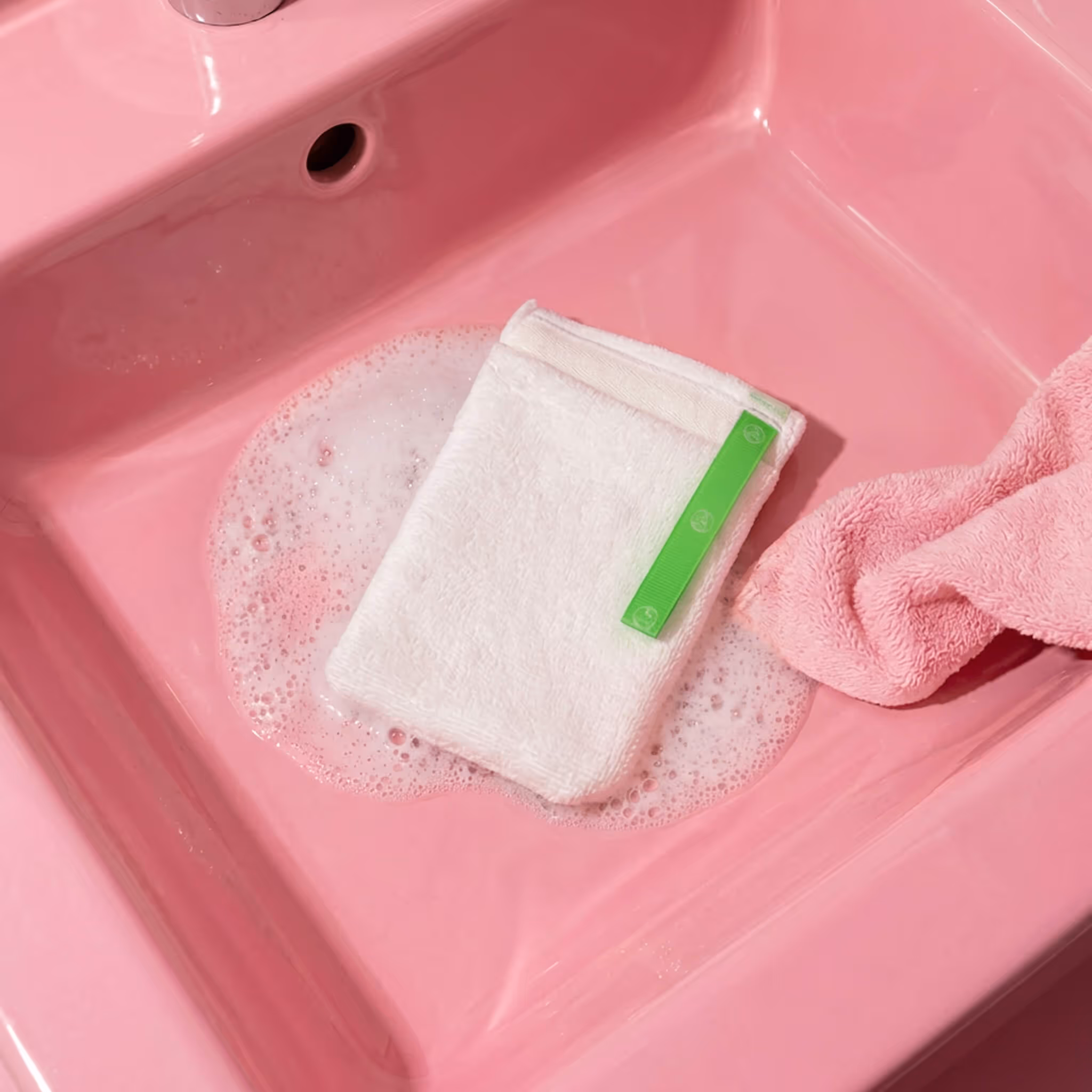 White AfterSpa Facial Scrubber mitt resting in pink sink with soap bubbles and towel nearby