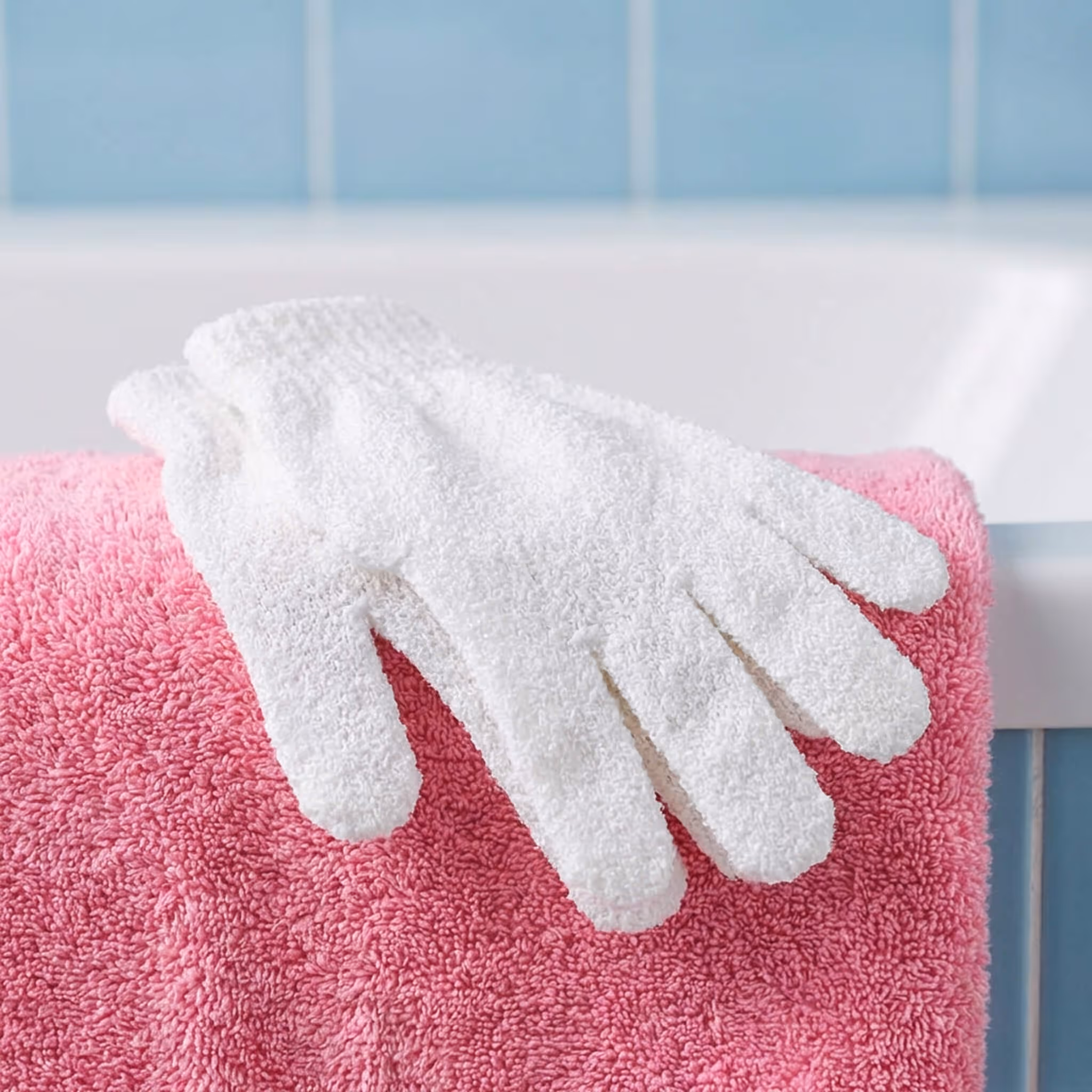 White exfoliating glove resting on folded pink towel beside bathtub with blue tile wall background.